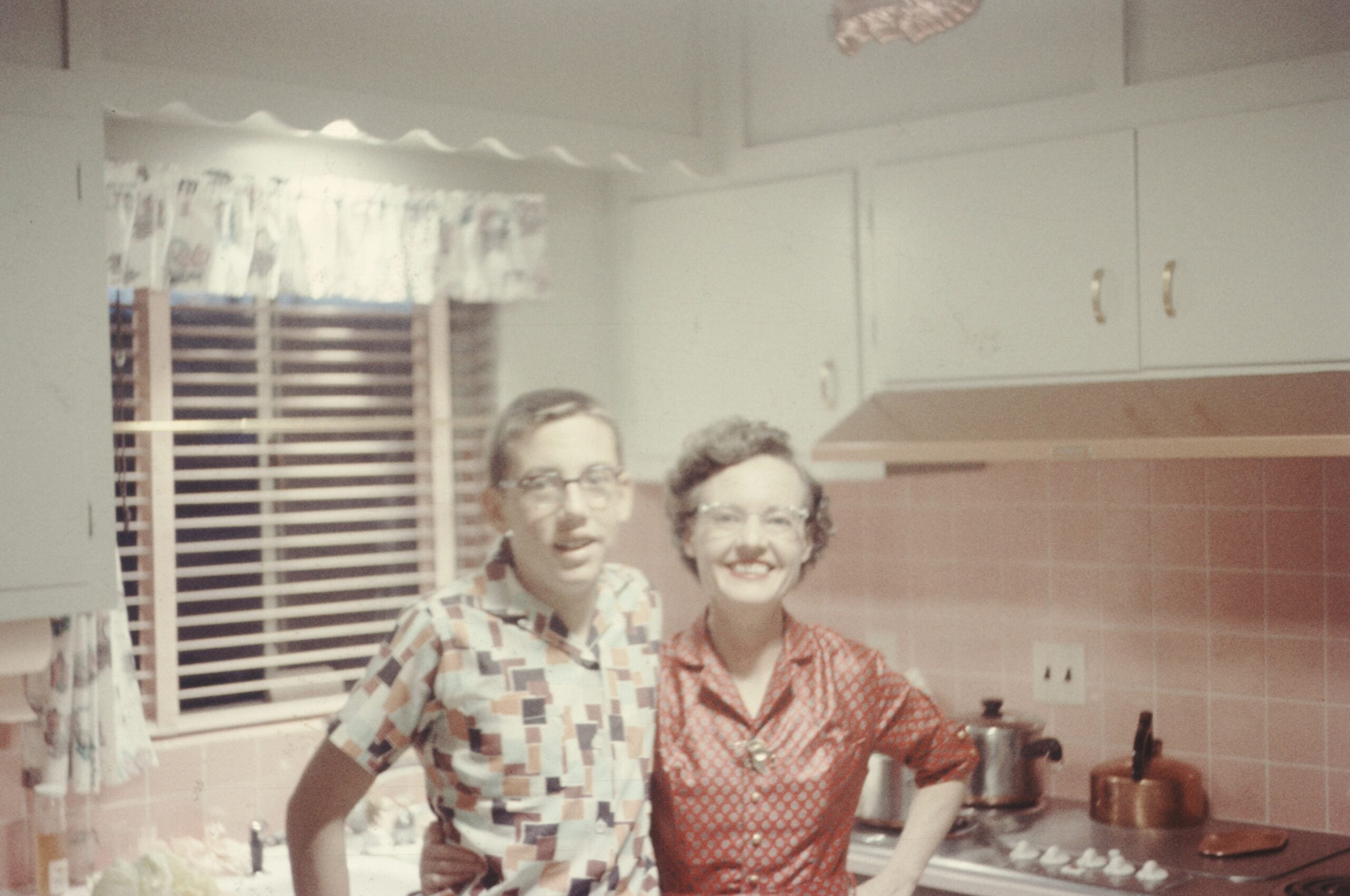 Two people smiling in a vintage kitchen with pink tiles, white cabinets, and patterned curtains. One wears a multicolored shirt, the other a red polka-dot dress. A stove and cookware are in the background.