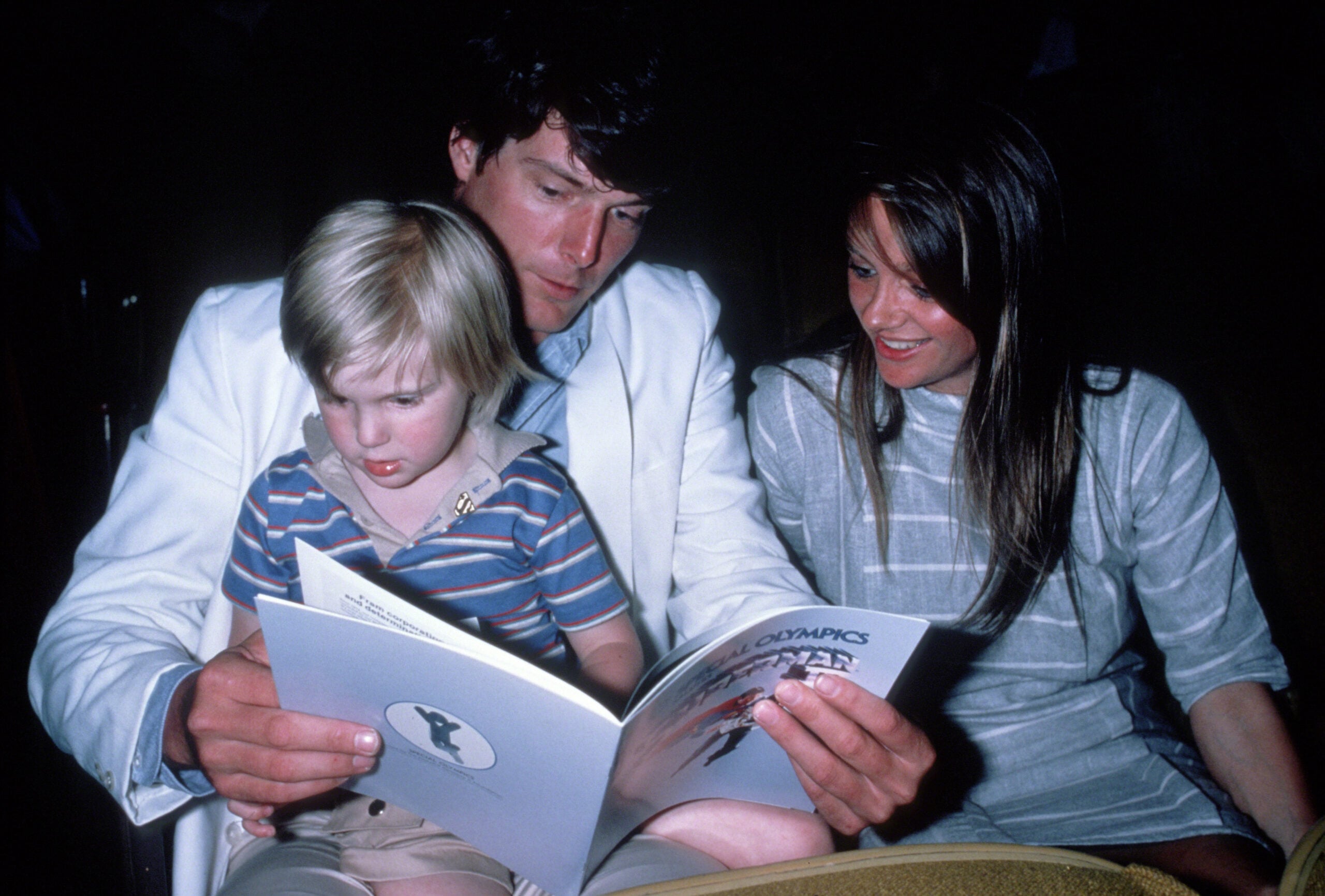 A man in a white suit reads a book with a young blond child on his lap, while a woman in a striped dress sits beside them, smiling.