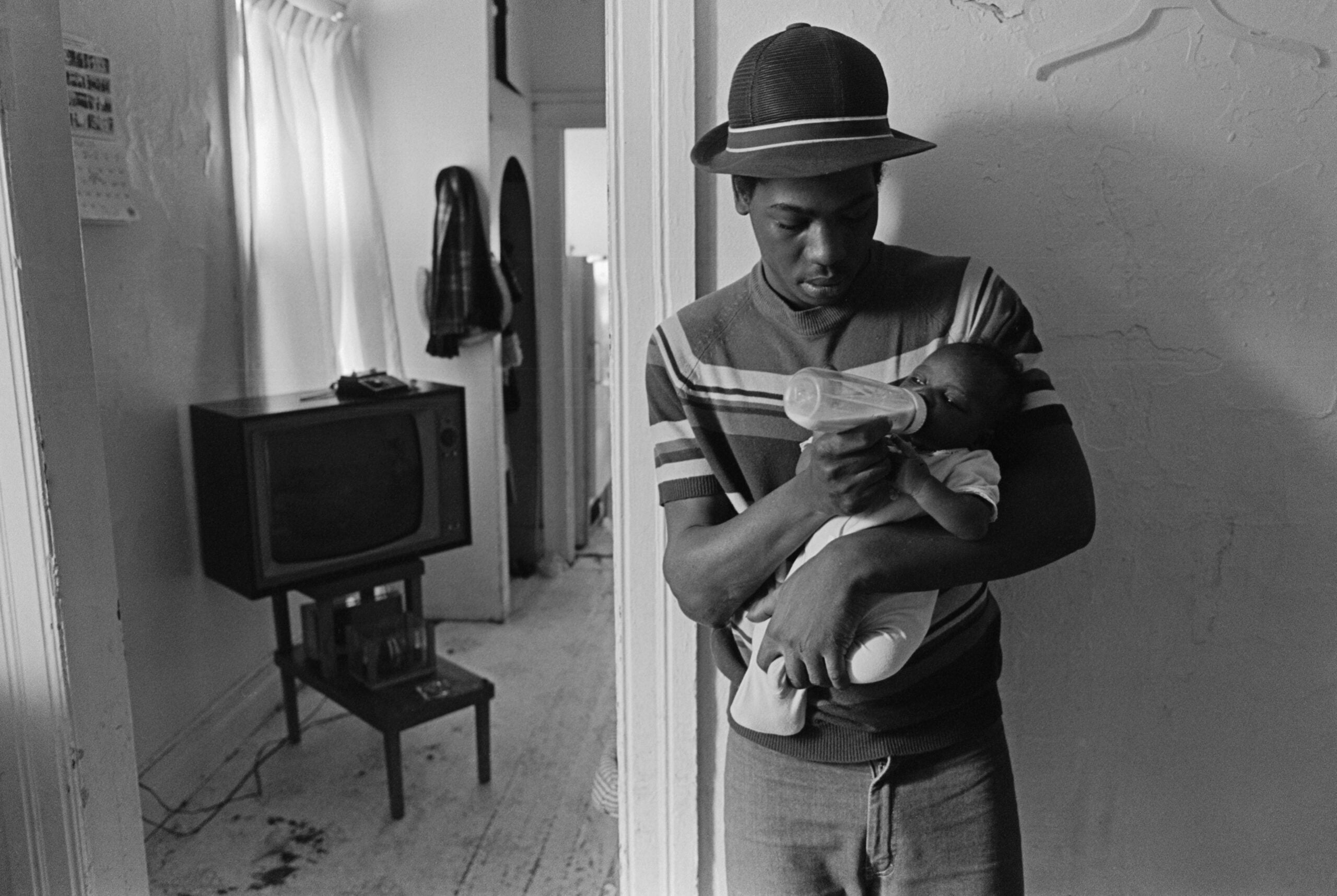 A man wearing a cap feeds a baby with a bottle in a modest room with a TV on a small table, a coat hangs on the wall, and light comes in through a curtained window.