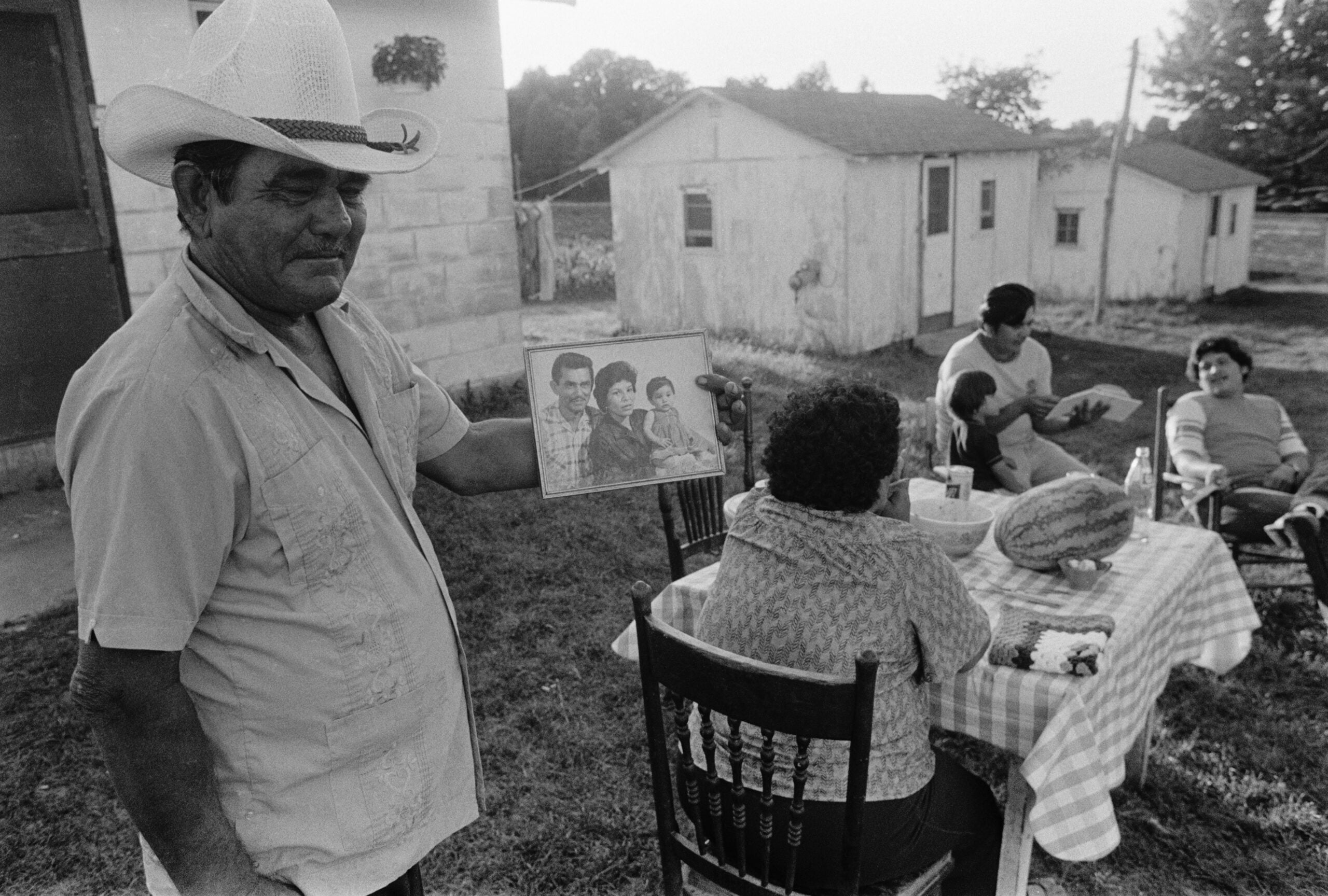 A man in a cowboy hat holds a family photo while others sit at a table outside, near small buildings, sharing food and conversation on a sunny day.