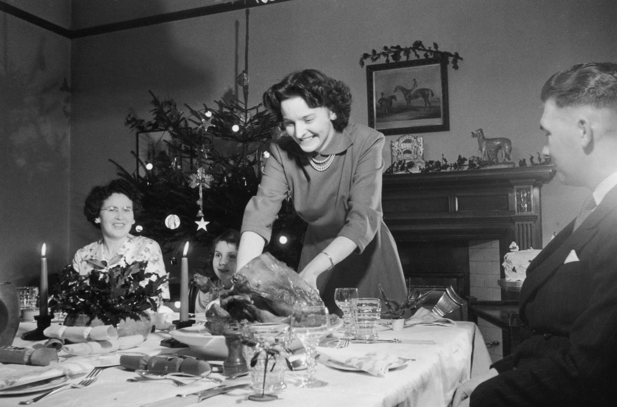 A woman carves a roast at a festive dinner table with a decorated Christmas tree in the background. Three other people sit around the table, smiling and enjoying the holiday meal together.