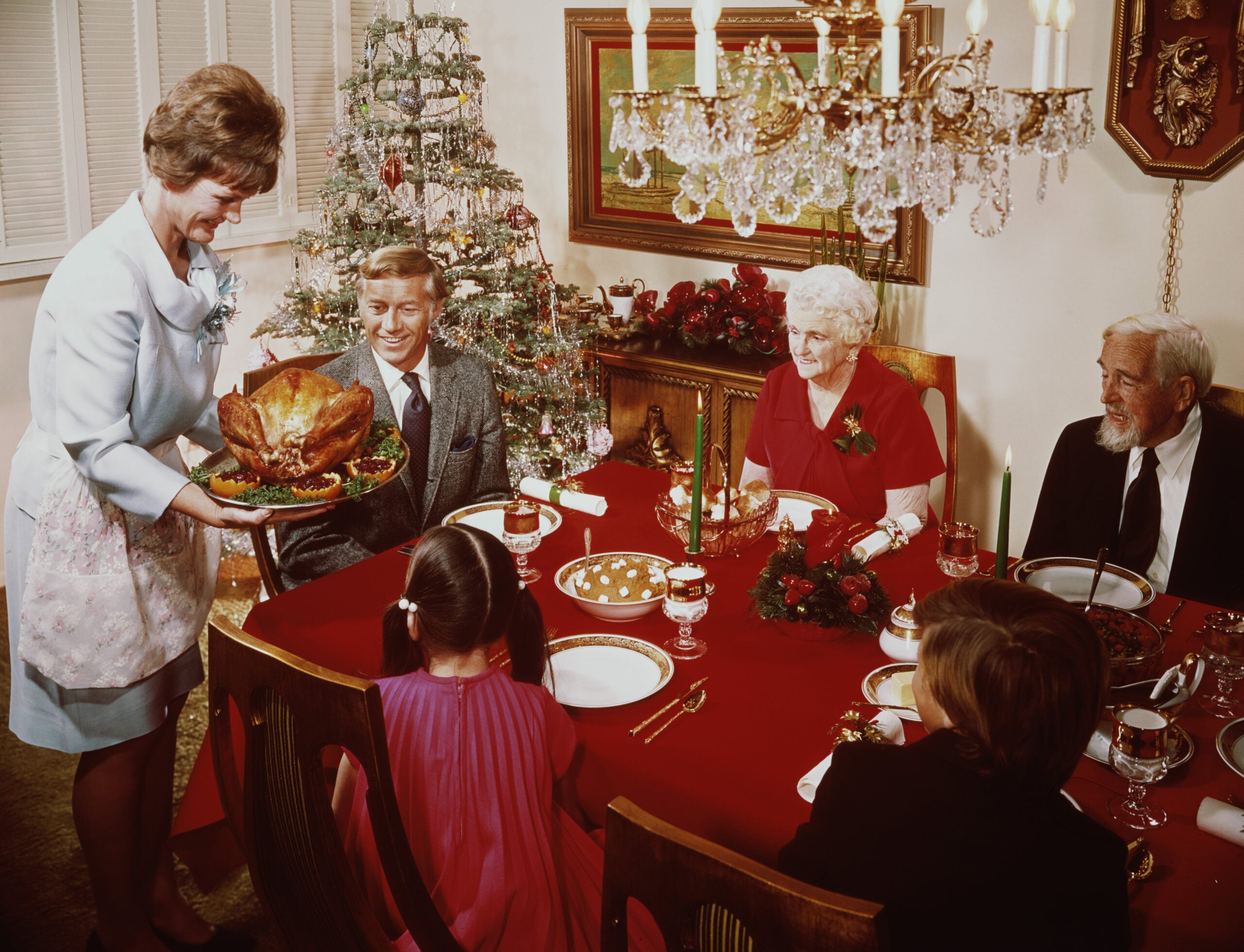 A woman serves a large roasted turkey to a smiling family gathered around a festive table, decorated for Christmas with a tree and holiday decor in the background.