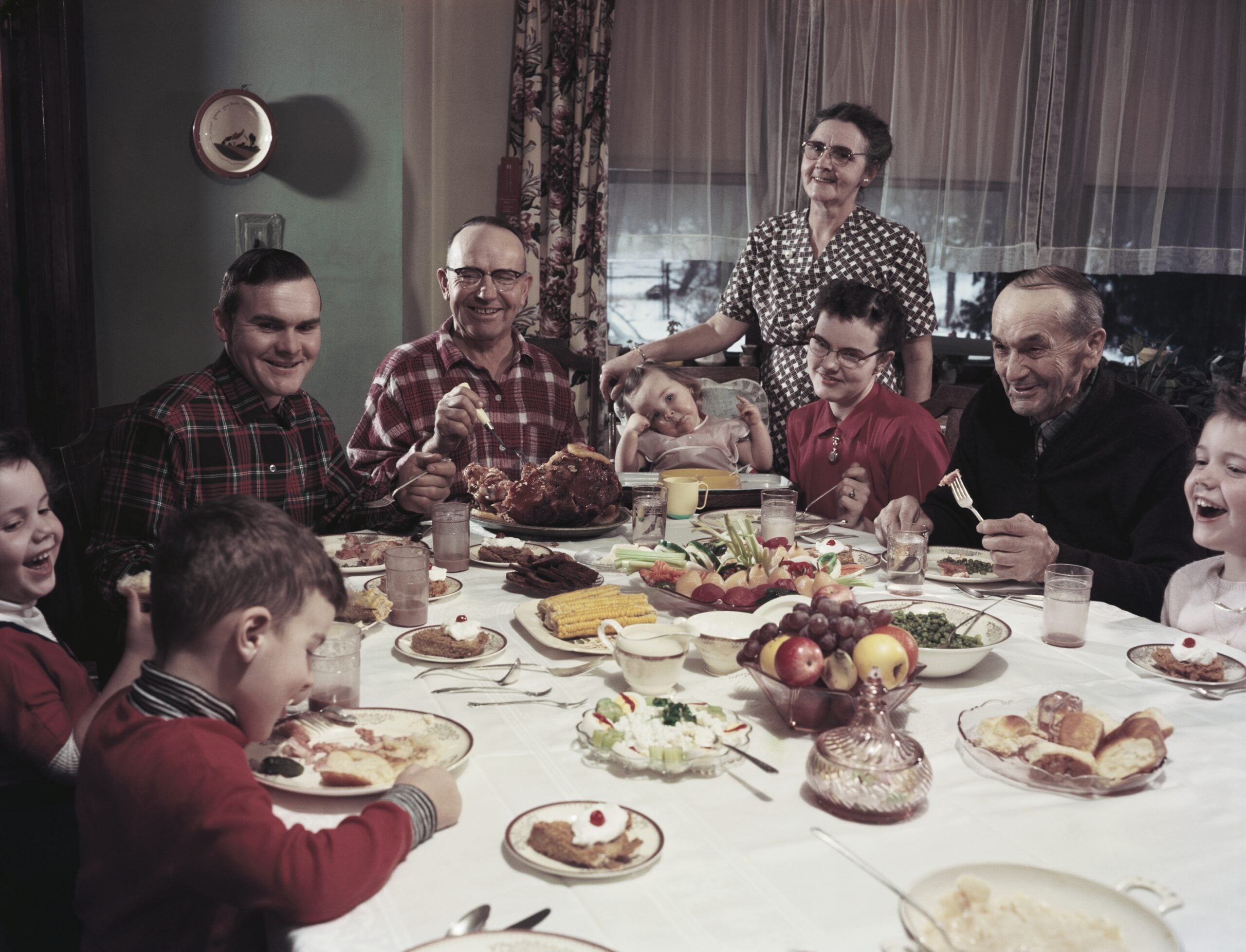 A family of nine, including children and elders, gathers around a dinner table set with various foods, smiling and laughing together in a warmly lit room with curtains and plants in the background.