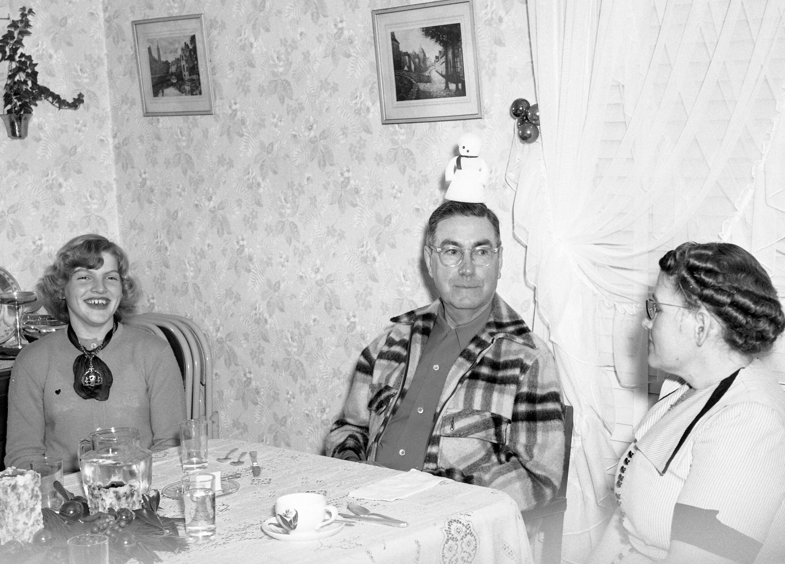 Three people sit at a dining table in a cozy room. The man in the center has a small snowman ornament on his head, making the woman on the left laugh. The woman on the right looks at him, smiling.