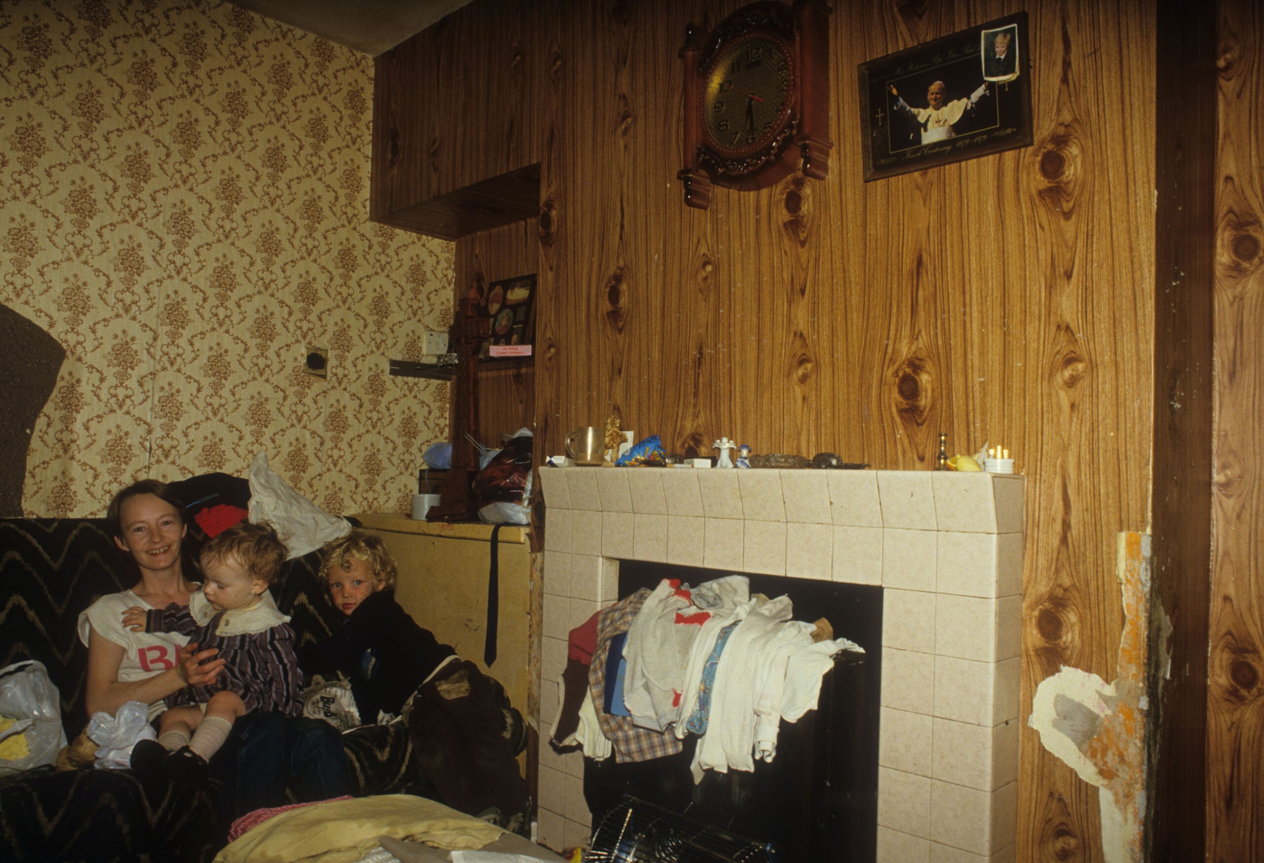 A woman with two children sits on a couch in a cluttered living room with patterned wallpaper, wood paneling, a fireplace, and clothes draped over the mantel. A clock and framed photo hang above the fireplace.