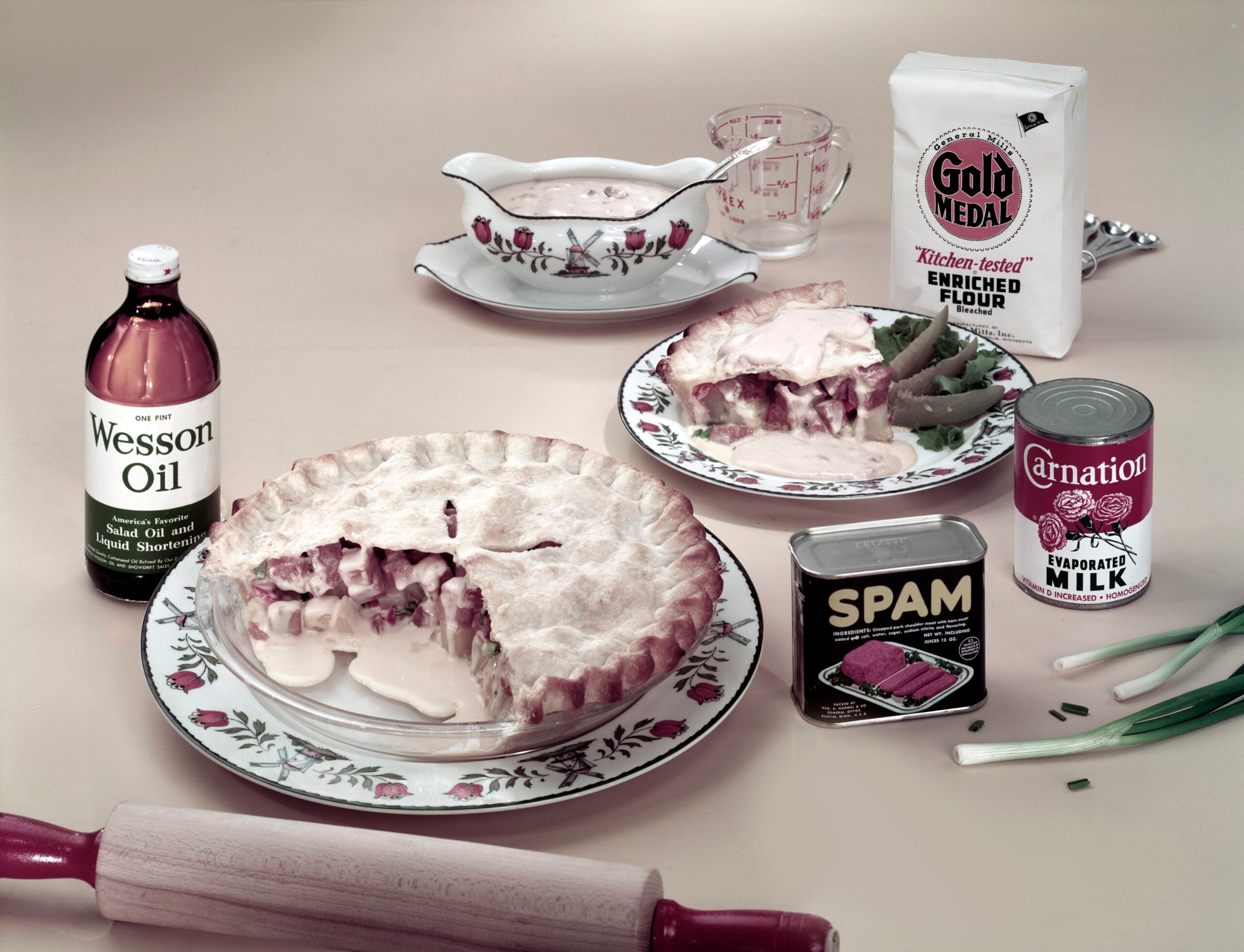 A table with a creamy pie on a plate, a slice served, surrounded by Spam, evaporated milk, flour, oil, green onions, a gravy boat, and a rolling pin, all on a floral-patterned set.