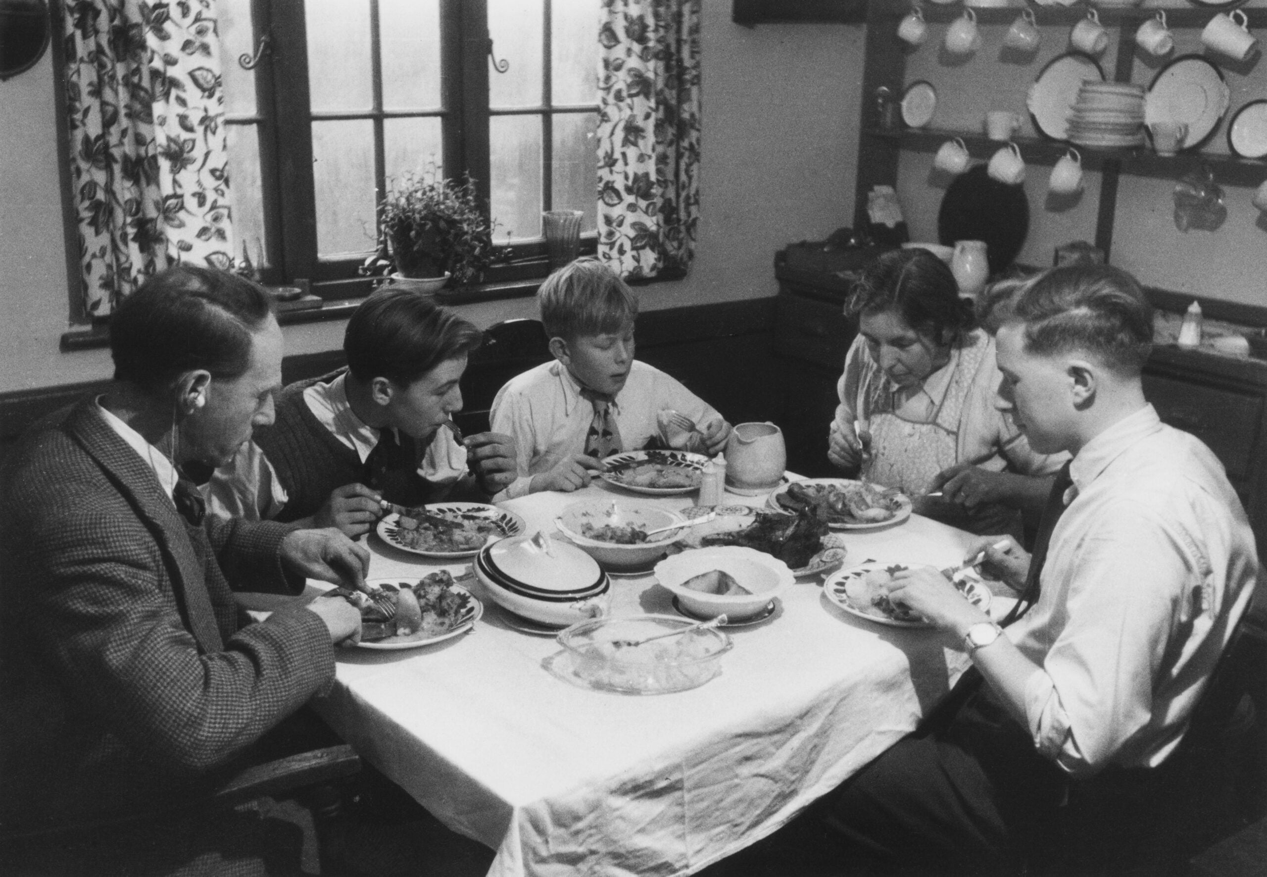 Five people sit around a table, eating a meal together in a cozy dining room with patterned curtains and shelves of dishes in the background.