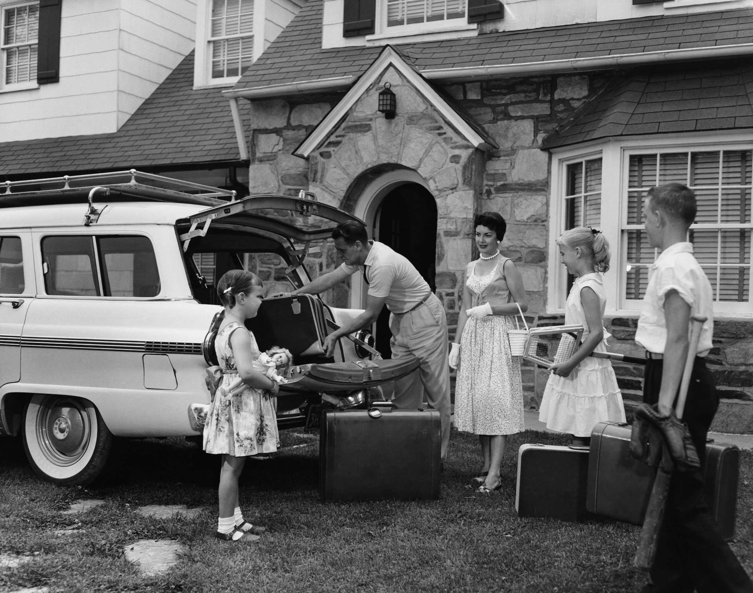 A family prepares for one of their classic family road trips as they pack suitcases into a station wagon outside their house. The father loads the car while the mother and three children stand nearby, ready for a trip. The photo appears to be from the 1950s or 1960s.