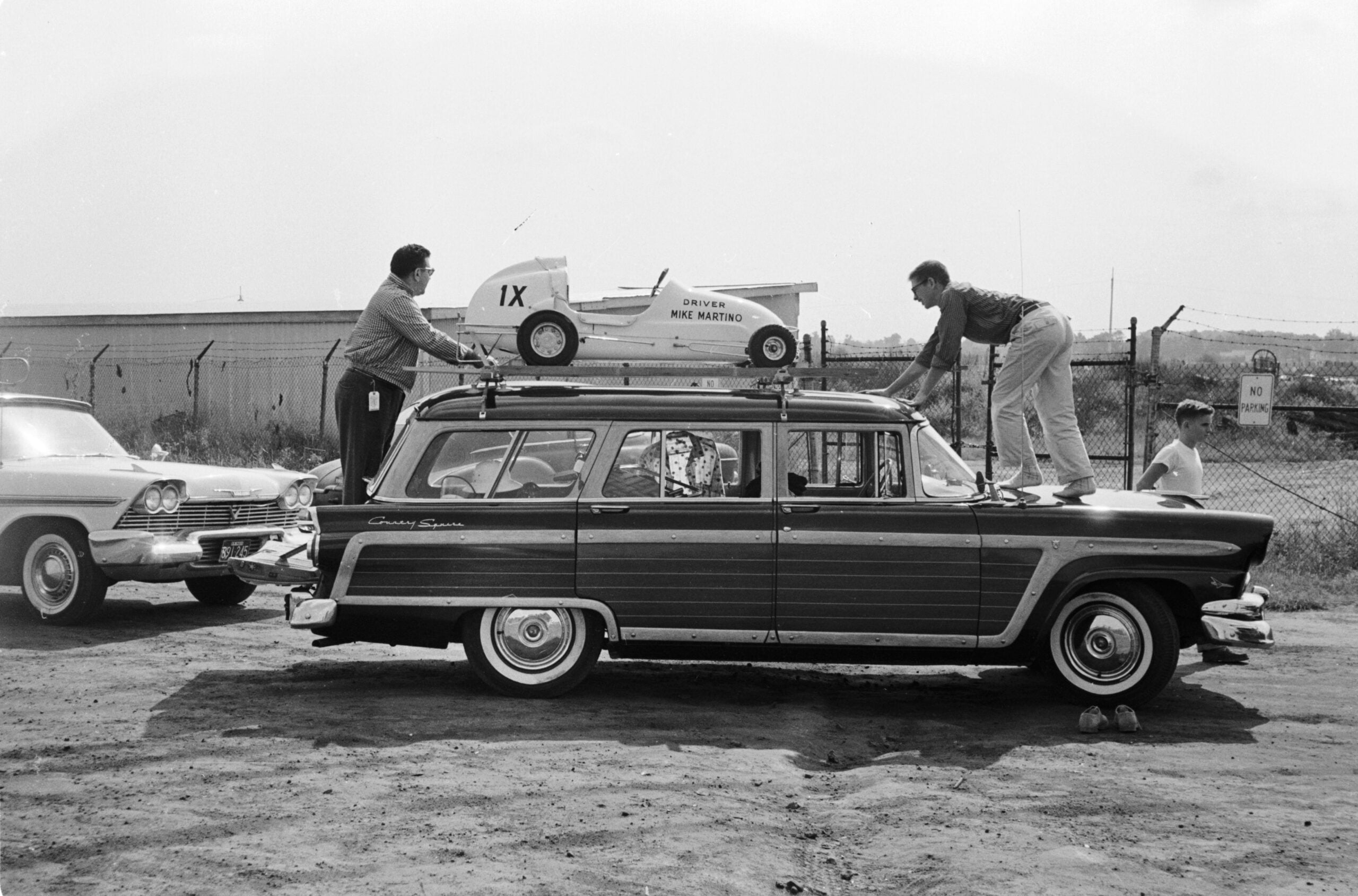 Two people secure a small race car labeled "Derby Mini Marathon" to the roof of a vintage station wagon, bringing to mind classic family road trips, while another person stands beside a nearby classic car in a dirt lot, with a chain-link fence in the background.