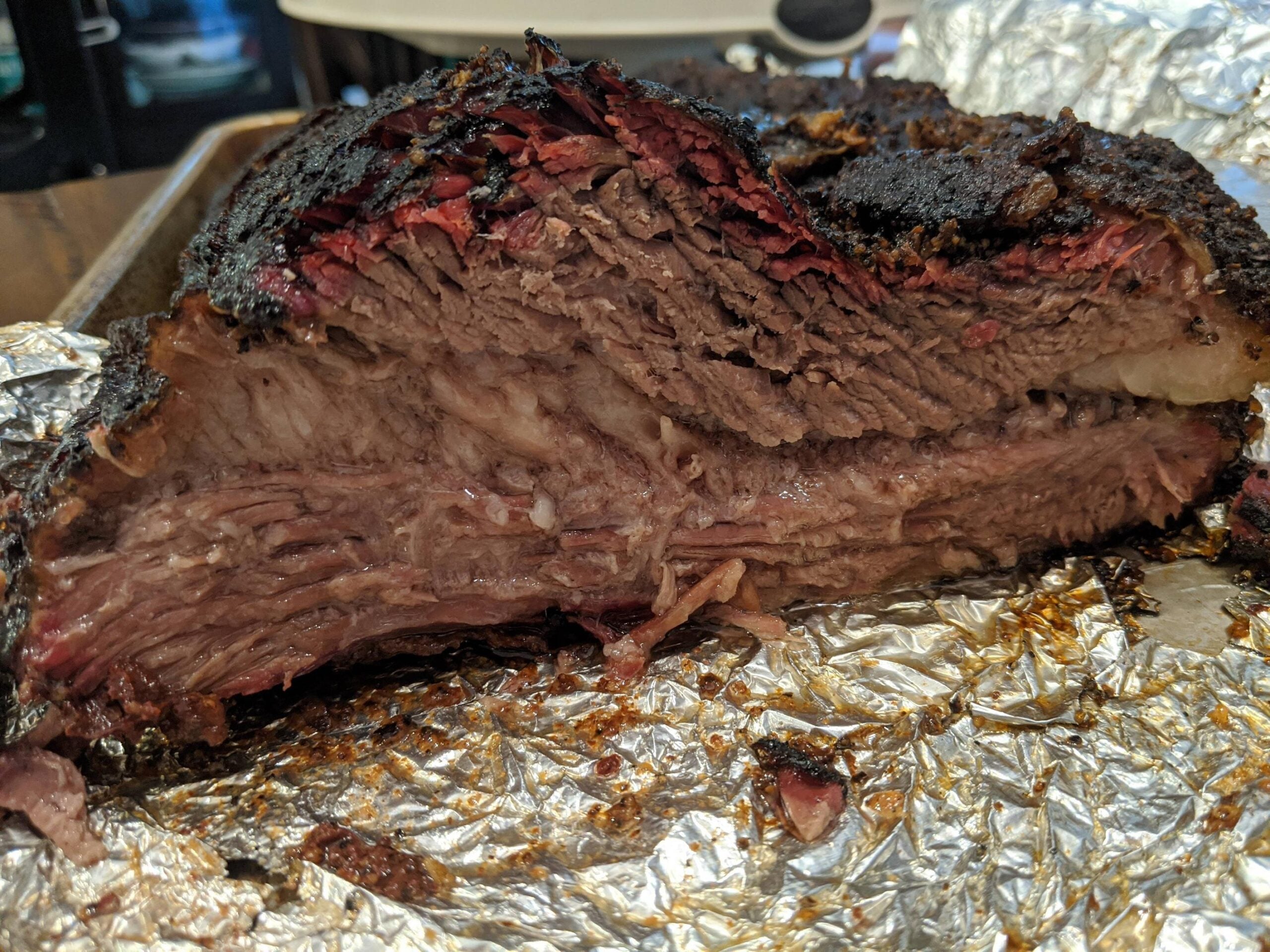 A close-up of a sliced smoked beef brisket, showing a dark bark, pink smoke ring, and juicy, tender meat, all resting on crinkled aluminum foil.