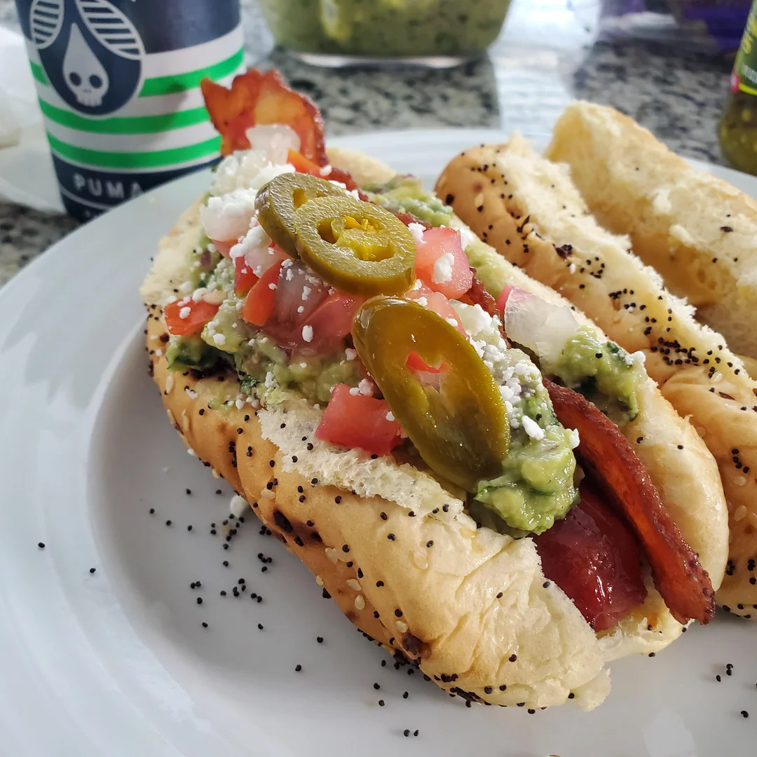 A poppy seed bun with a hot dog, bacon, guacamole, diced tomatoes, onions, queso fresco, and jalapeño slices, served on a white plate. A drink cup and another hot dog are in the background.
