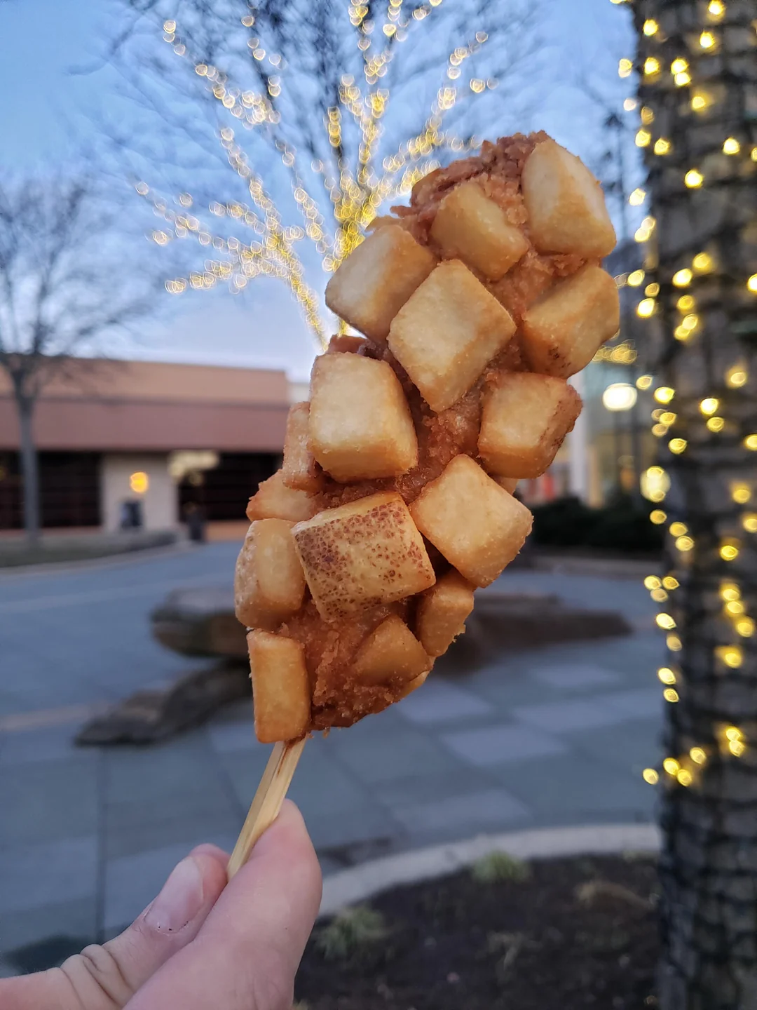 A hand holds a Korean corn dog covered in crispy, golden potato cubes. The background shows trees with glowing string lights and an outdoor plaza at dusk.