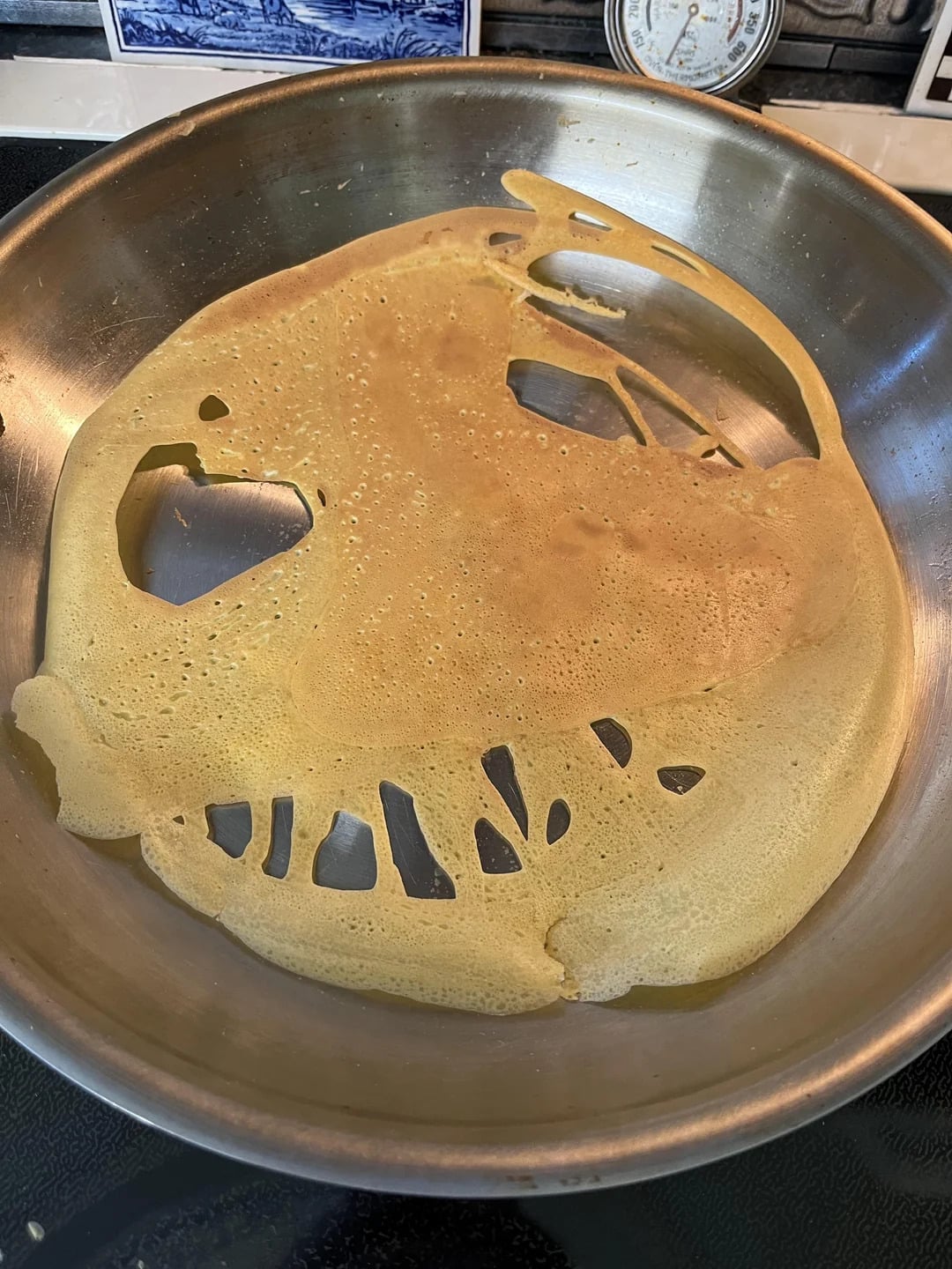 A thin pancake cooking in a skillet with holes, resembling a fish shape with an open mouth and gills. A stove and thermometer are visible in the background.