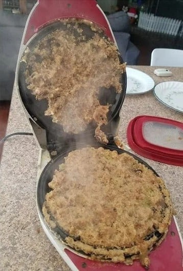 A waffle maker opened to reveal a messy, overcooked waffle stuck to both plates, with steam rising and crumbs scattered on the counter nearby.