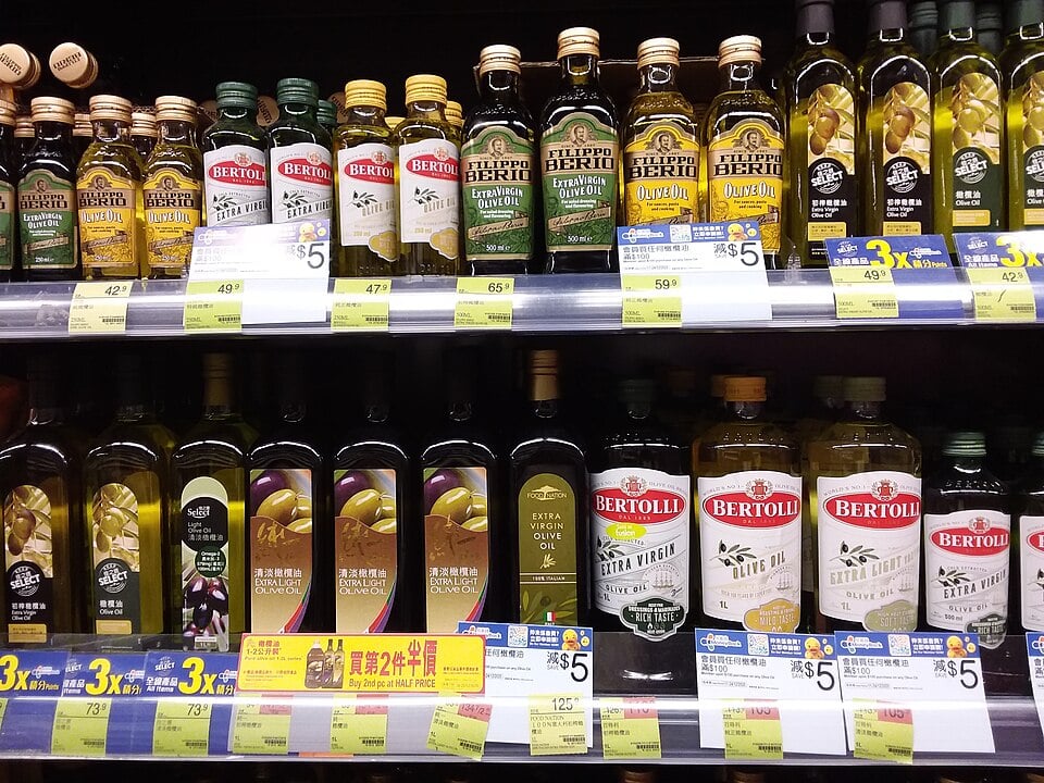 Bottles of olive oil from various brands, including Filippo Berio and Bertolli, are displayed on supermarket shelves with yellow price tags and promotion signs.