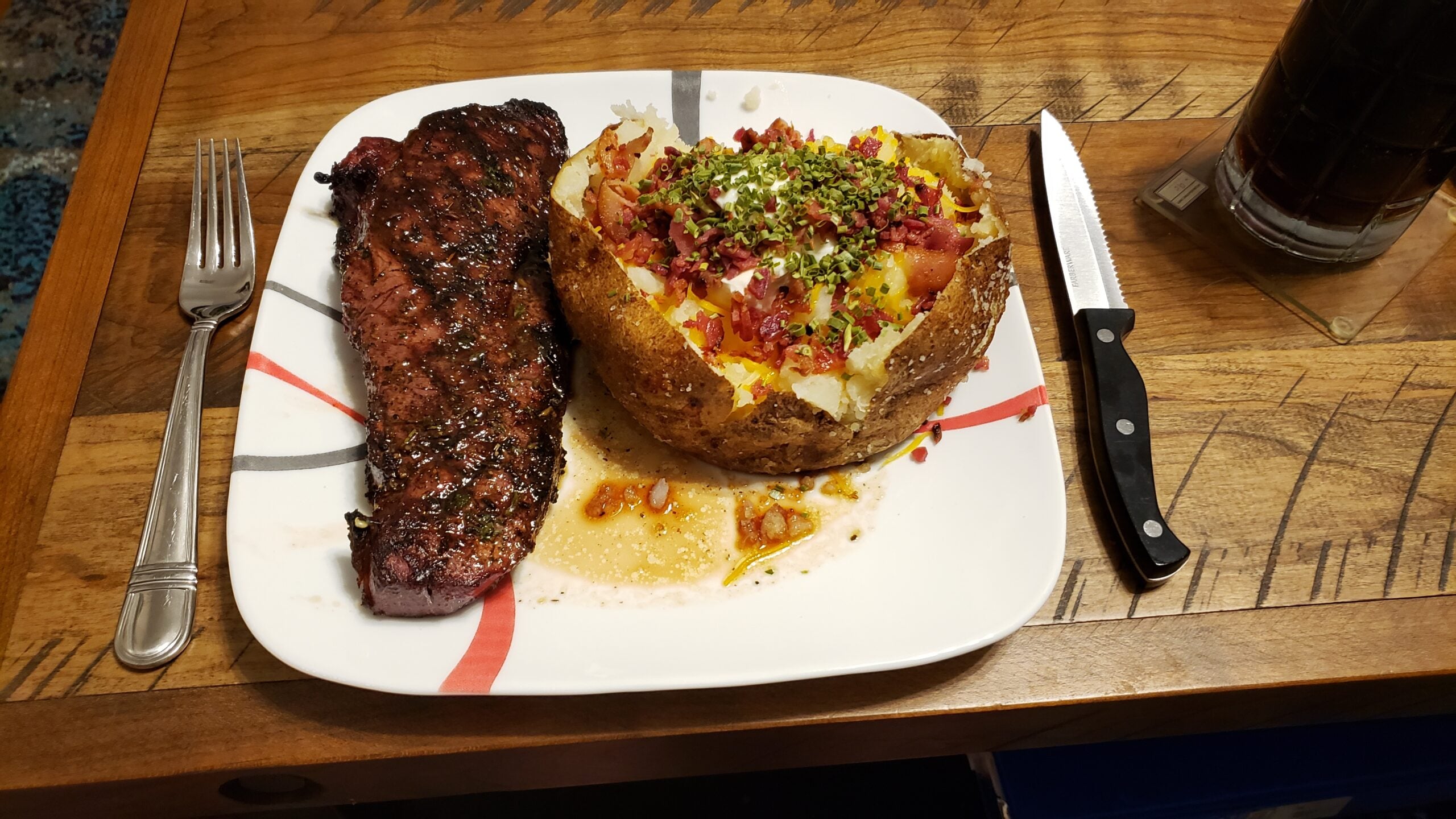 Grilled steak with a loaded baked potato topped with cheese, bacon bits, chives, and sour cream on a plate, with a knife, fork, and a glass of dark soda on a wooden table.