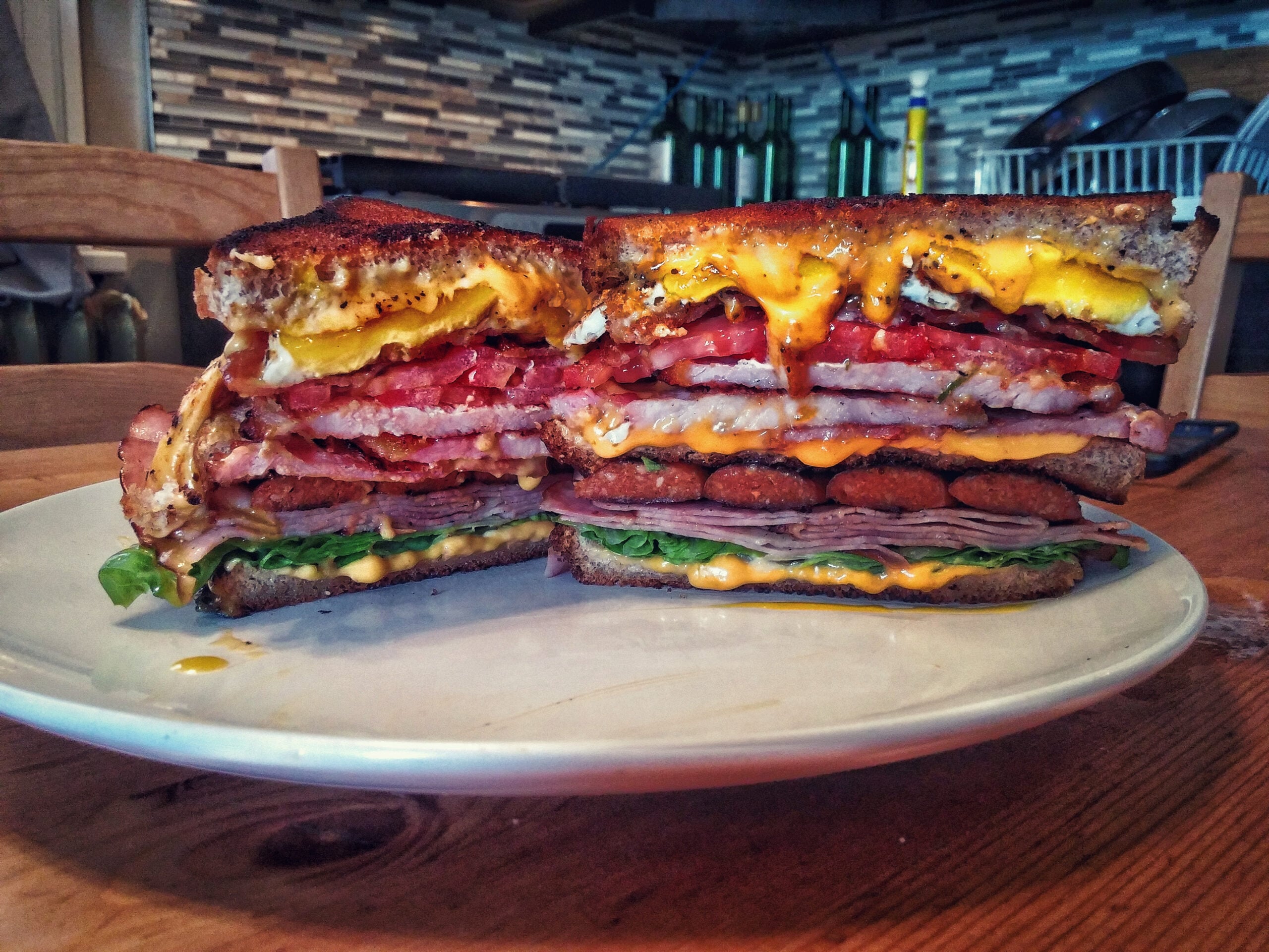 A thick, stacked sandwich on toasted bread with layers of egg, bacon, sausage, ham, lettuce, and melted cheese, served on a white plate on a wooden table in a kitchen.