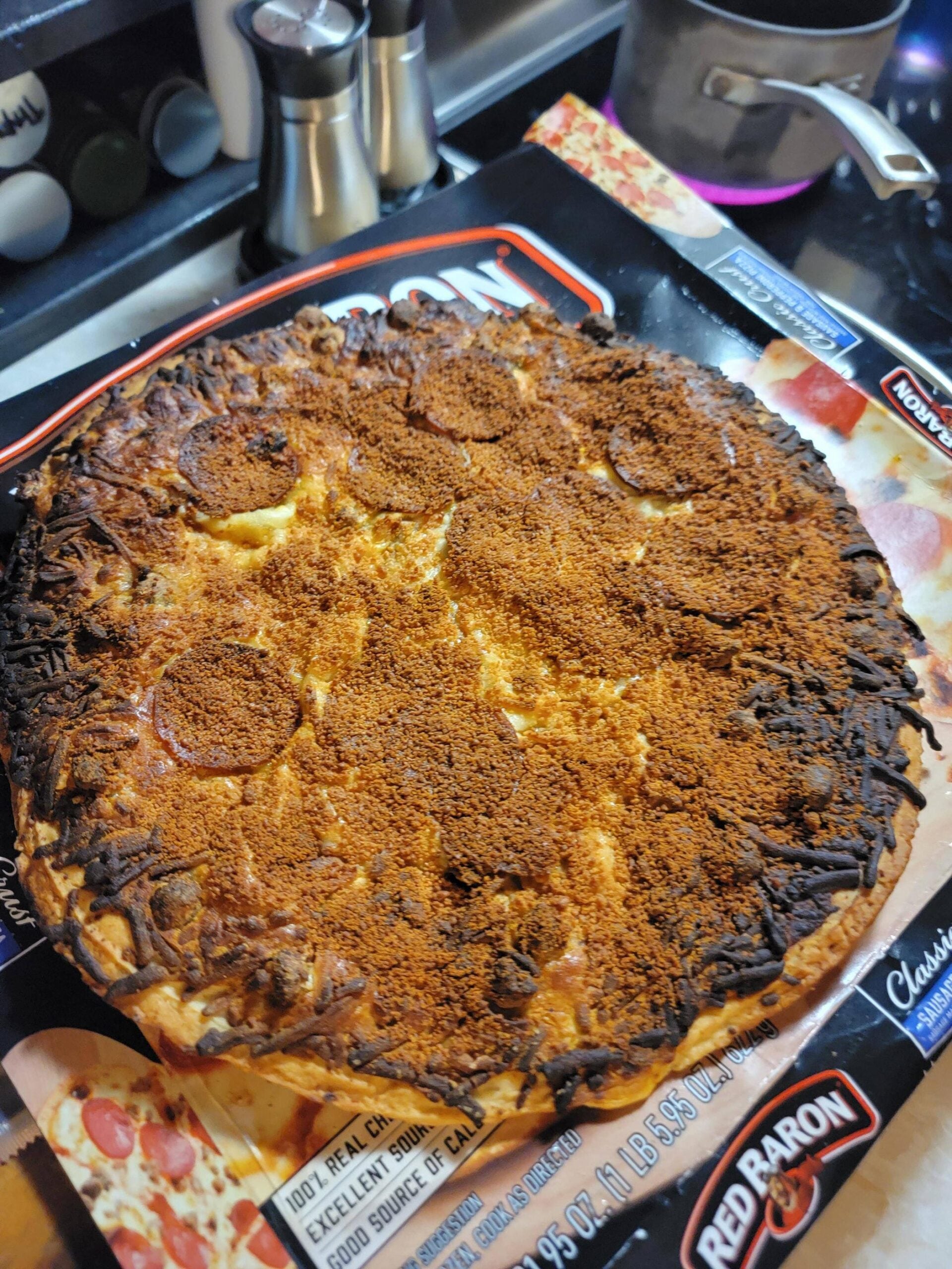 A frozen Red Baron pizza is overcooked, with a very burnt top and edges, still on its packaging, sitting on a kitchen counter near spices, salt and pepper shakers, and a pot on the stove.