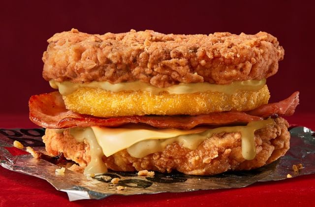 Two crispy fried chicken fillets sandwiching cheese, bacon, a hash brown, and sauce, with no traditional bun, against a red background.