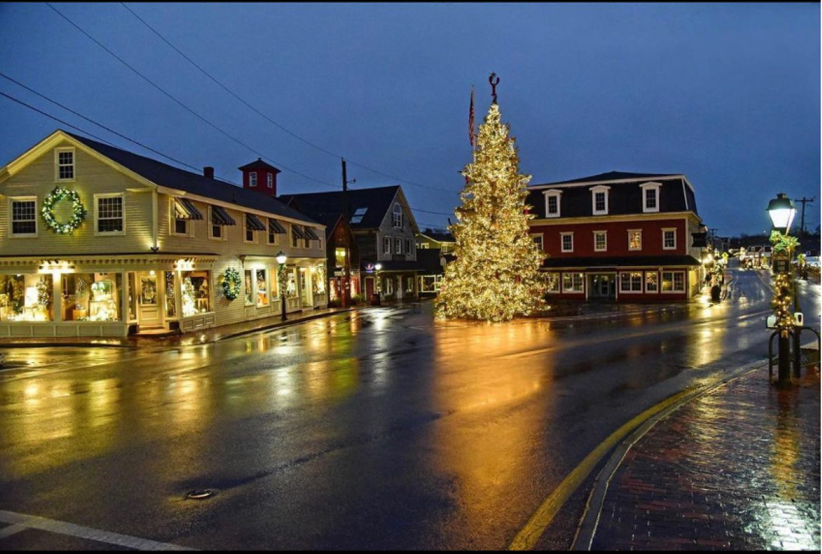 A quiet, wet street corner at night, featuring lit storefronts, holiday wreaths, and a large Christmas tree adorned with lights in the town square—capturing the magical charm found in some of the best Christmas towns in America.