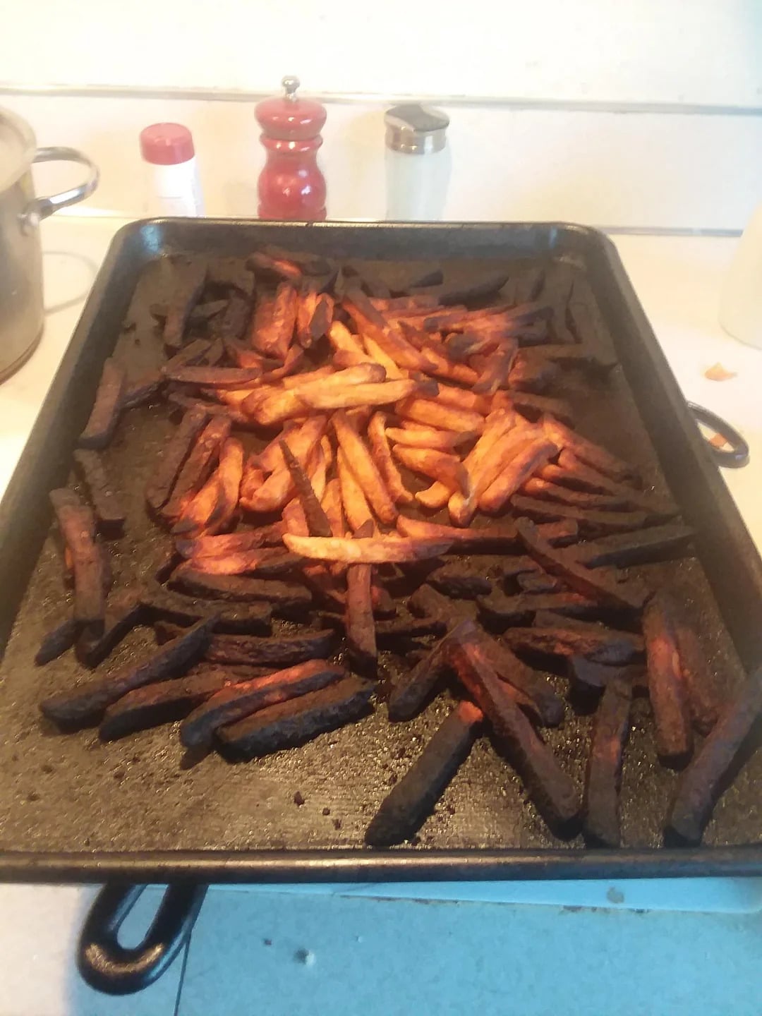 A baking tray filled with burnt, overcooked French fries sits on a kitchen counter. Cooking pots, a red pepper grinder, and a salt shaker are visible in the background.