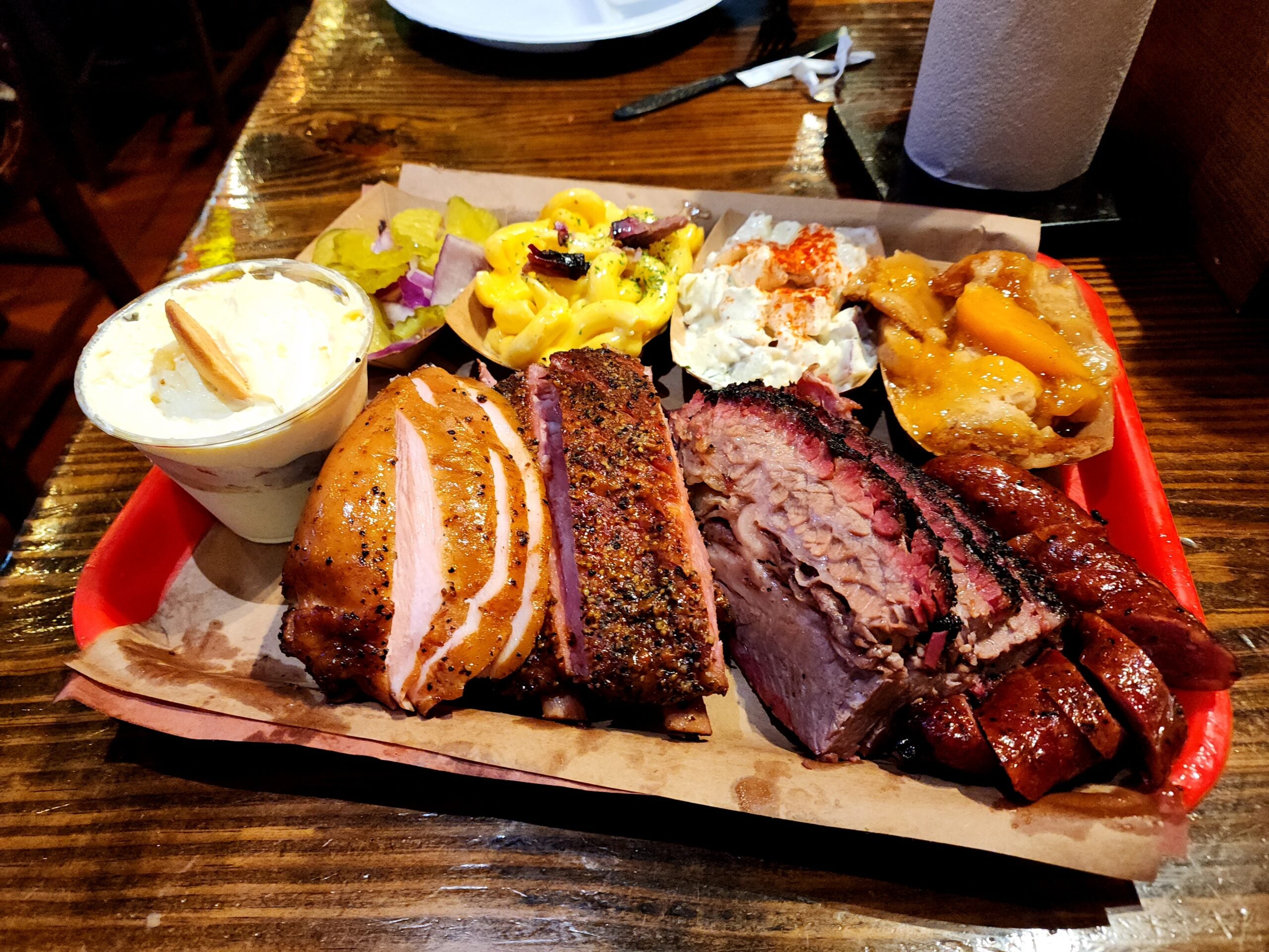 A barbecue platter with ribs, sausage, brisket, and chicken, served with sides of macaroni and cheese, coleslaw, potato salad, pickles, onions, and baked beans on a red tray on a wooden table.