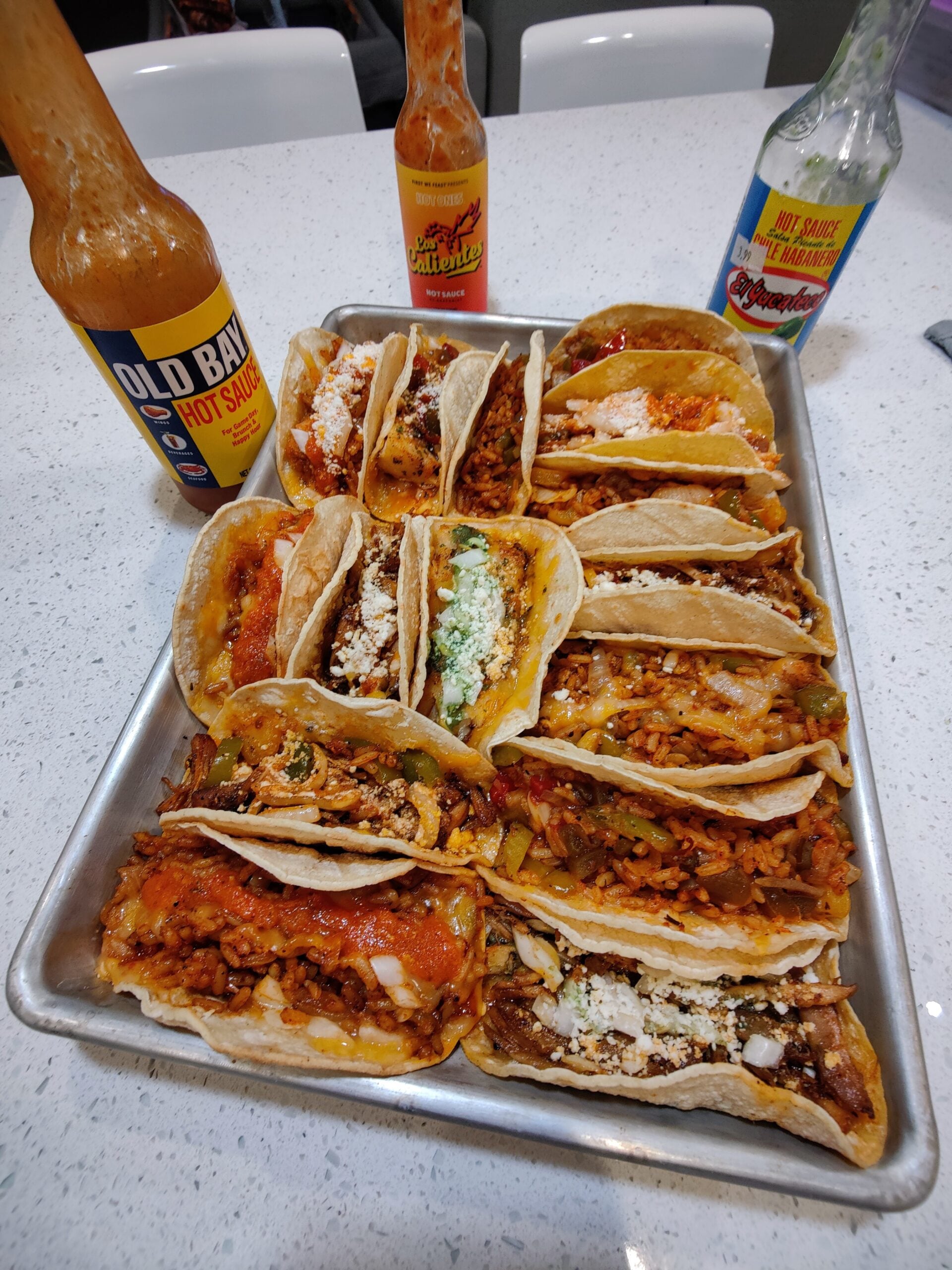 A tray of assorted tacos filled with various meats, cheeses, and toppings sits on a counter, perfect for the best BBQ cookouts this summer. Three bottles of hot sauce are placed behind the tray, suggesting a flavorful taco meal everyone will enjoy.
