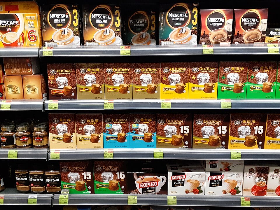 Supermarket shelf displaying various brands of instant coffee, including Nescafé, Old Town, and Kopiko, arranged in colorful boxes and jars with labels facing forward.