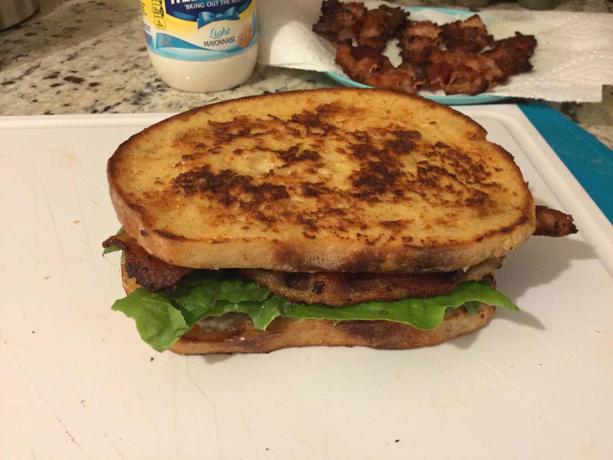 A toasted sandwich with lettuce, bacon, and mayonnaise sits on a white cutting board. In the background, there is a jar of mayonnaise and some cooked bacon on paper towels.