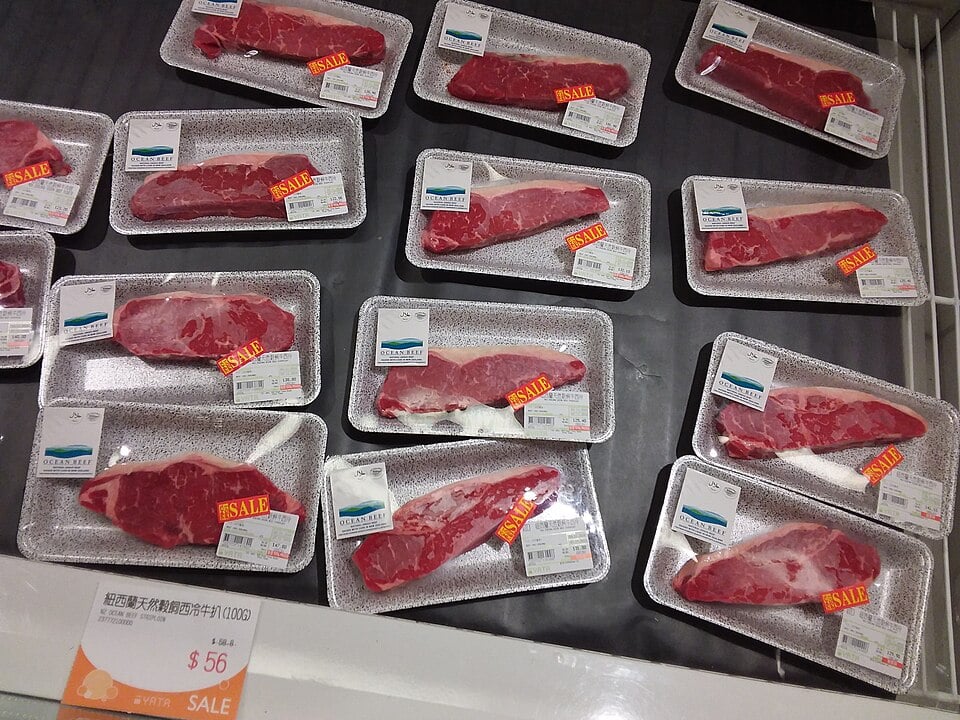 Several packages of raw beef cuts in plastic trays with sale stickers are displayed in a refrigerated supermarket case. A price tag in the foreground reads "$56" in red and white.