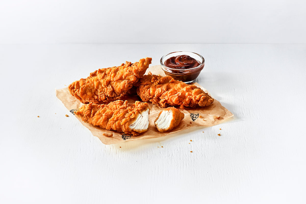 Three crispy fried chicken tenders, reminiscent of those served at popular fried chicken chains, are placed on parchment paper with a small cup of dark dipping sauce next to them. One tender is cut open, showing the white meat inside. The background is a clean white surface.