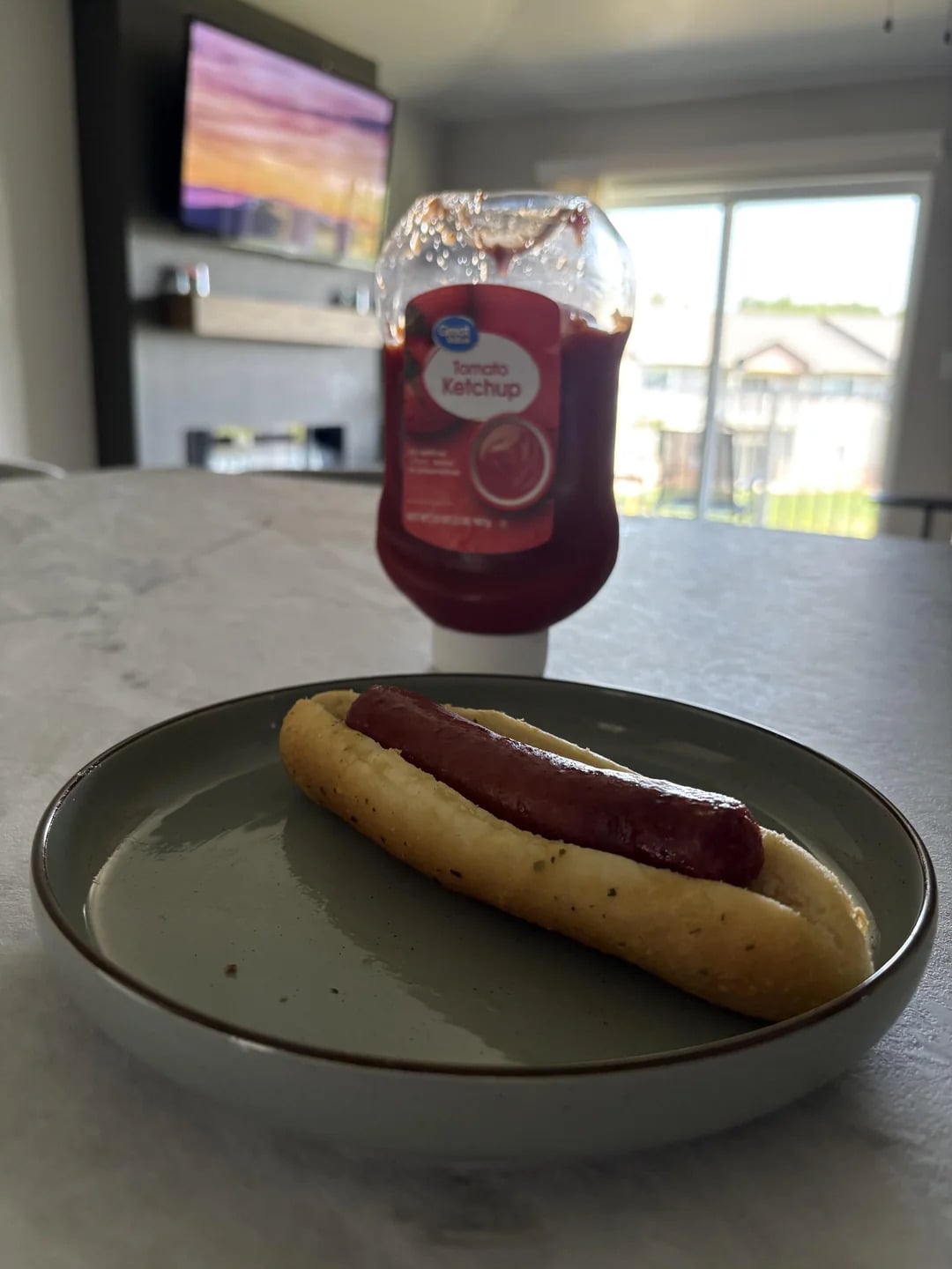 A plain hot dog in a bun sits on a green plate on a table. Behind it, there is a bottle of tomato ketchup and a TV showing a colorful screen in a bright living room.