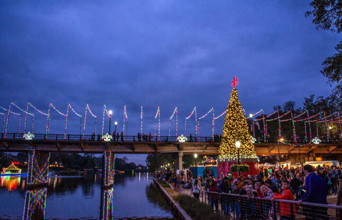 A crowd gathers by a river near a large Christmas tree and a bridge decorated with colorful holiday lights at dusk, capturing the festive charm that makes this one of the best Christmas towns in America.