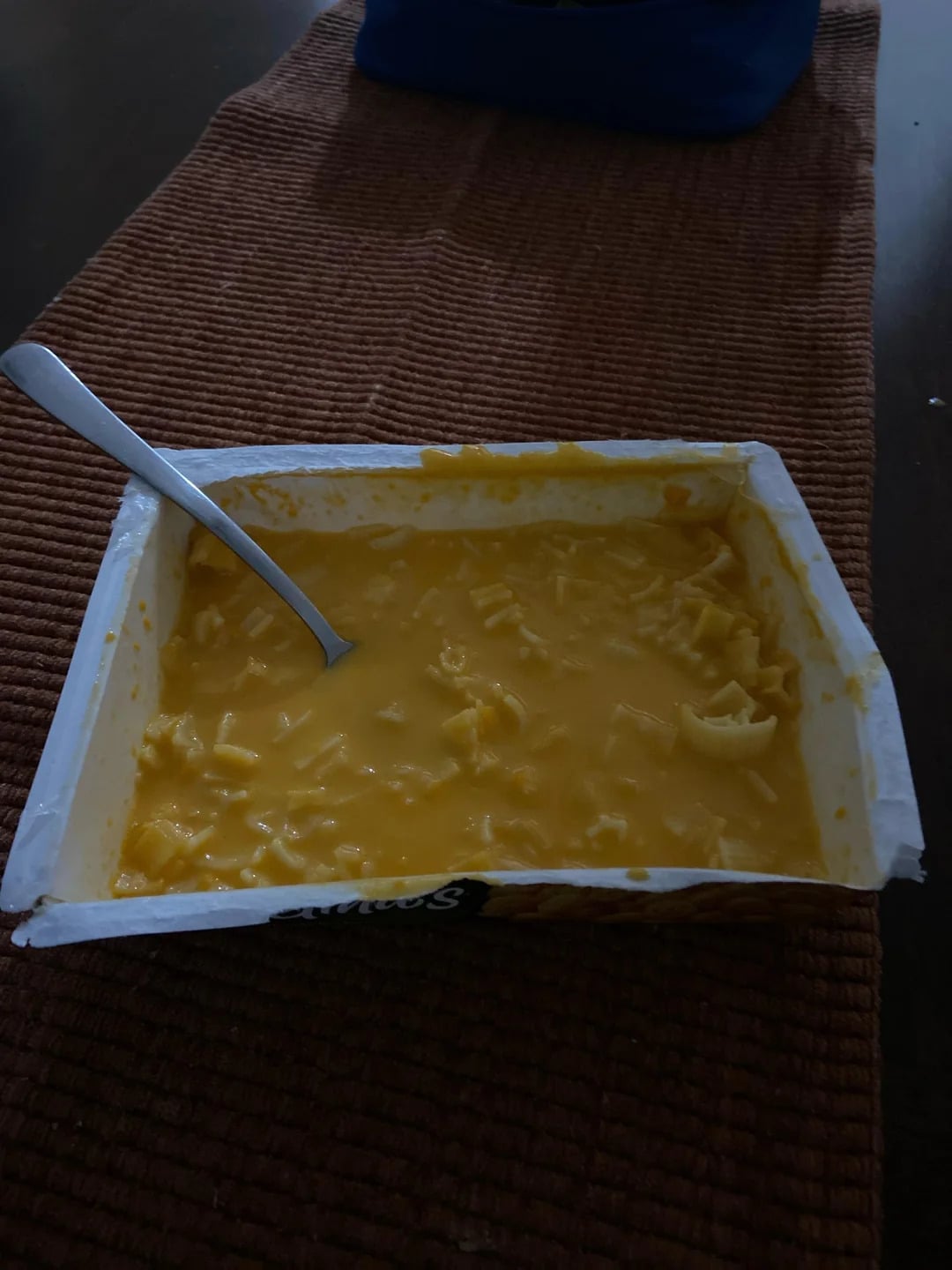 A rectangular container of macaroni and cheese with a spoon in it sits on a textured brown table runner.