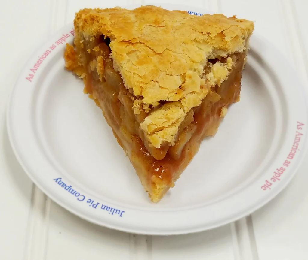 A slice of apple pie with a golden, flaky crust sits on a white plate. The apple pies filling is visible, showing baked apple slices and syrup. The plate has blue and red text around the rim.