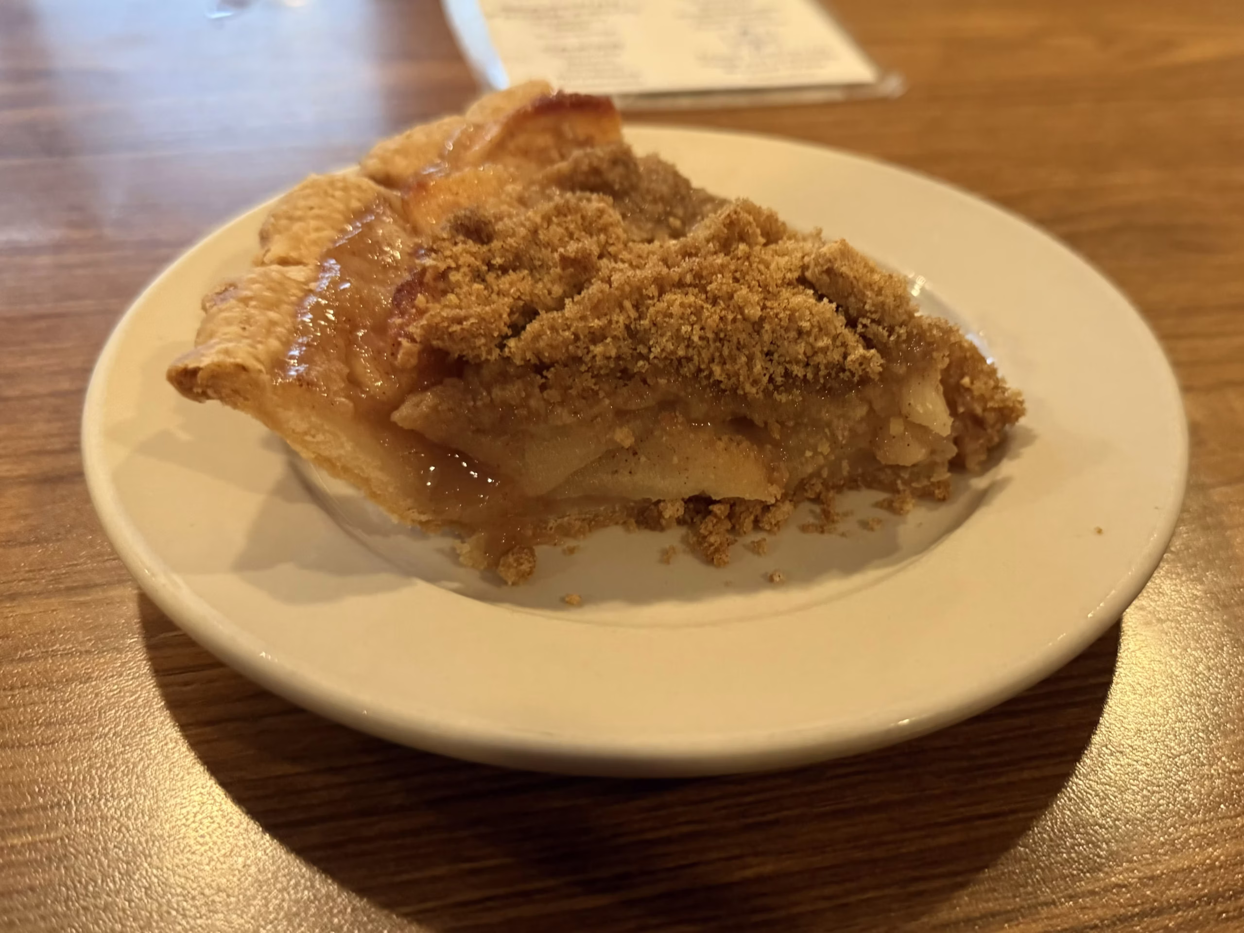 A slice of crumb-topped apple pie, one of the classic apple pies, sits on a white plate on a wooden table, with a blurred piece of paper in the background.