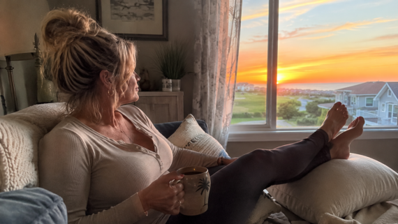 A woman with blonde hair in a messy bun relaxes on a cozy couch, mug in hand, gazing at a sunset over houses&mdash;enjoying the benefits of renting as warm light fills her softly decorated space.