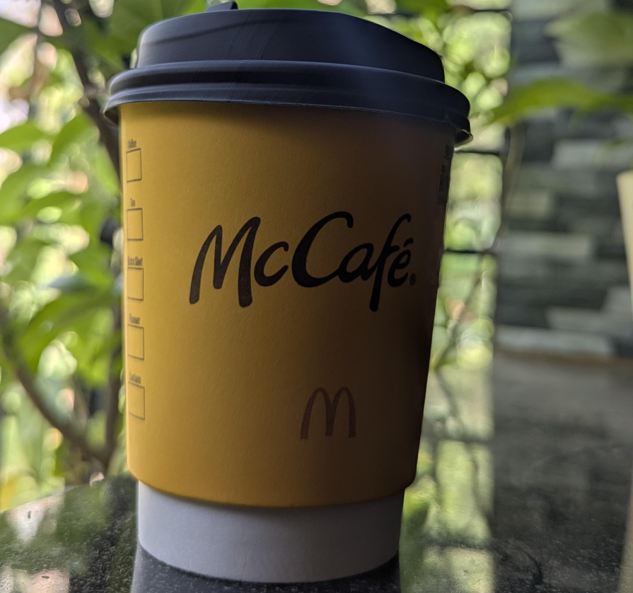 A yellow McCafé coffee cup with a black lid sits on a reflective surface, showcasing the distinctive style of fast food coffees, with green leaves and a blurred outdoor background behind it.