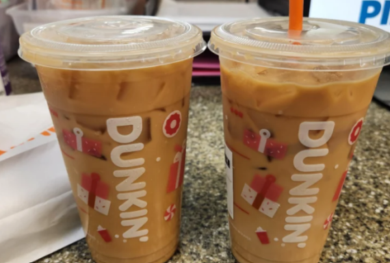 Two large fast food coffees from Dunkin&rsquo; sit on a granite countertop, each with a plastic lid and straw. The cups display the Dunkin&rsquo; logo and playful red graphics, while other items are blurred in the background.