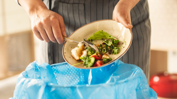 A person scrapes leftover salad into a trash bin lined with a blue plastic bag, mindful that reducing food waste can help manage household expenses. They're wearing a striped apron, and the salad consists of greens and vegetables.