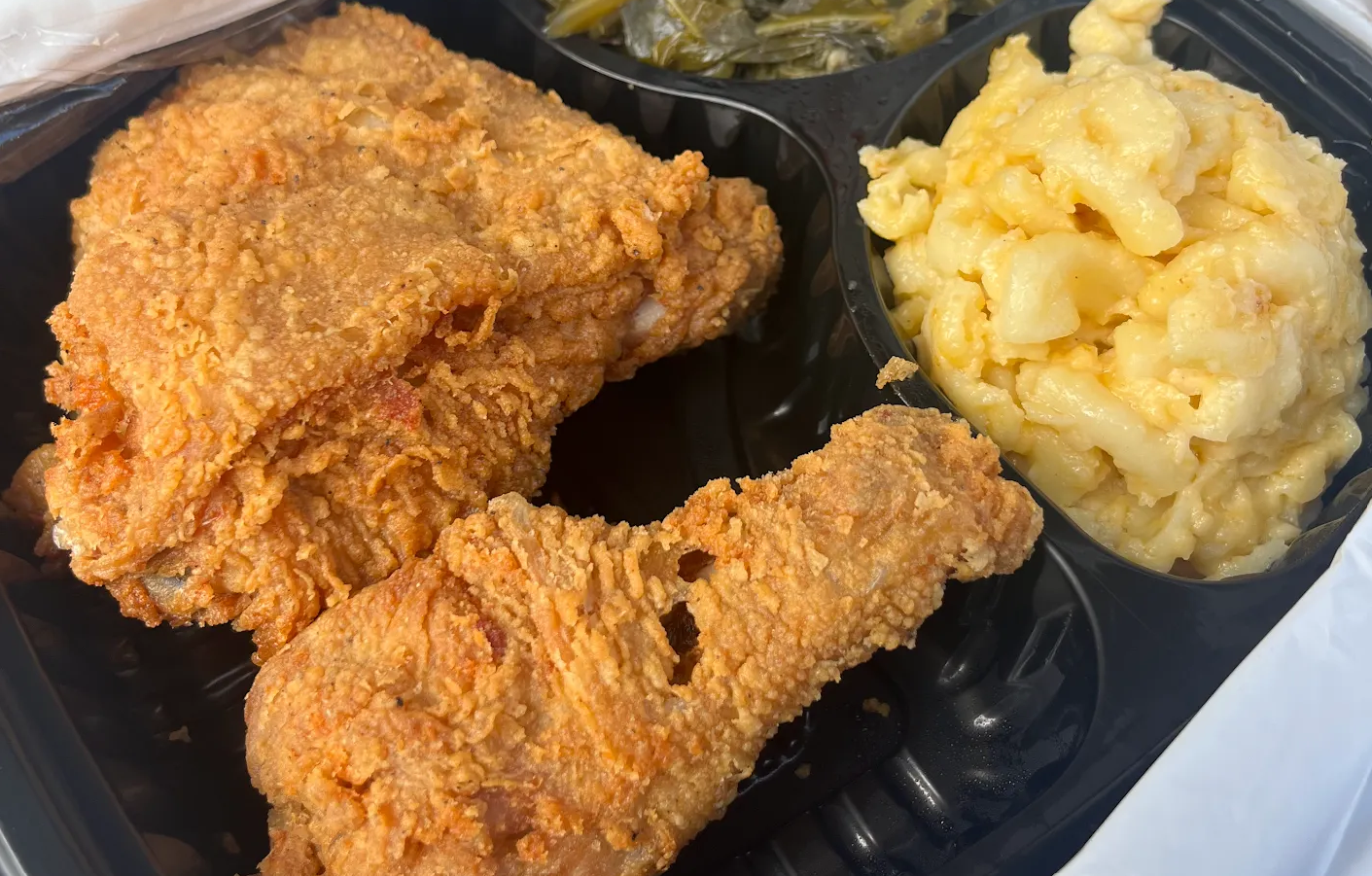 A black tray featuring one of our signature fried chicken dishes, served with creamy macaroni and cheese and a side of cooked greens.