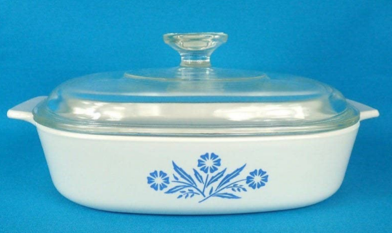 A white ceramic casserole dish with handles and a clear glass lid, featuring a blue floral pattern on the front, set against a blue background&mdash;perfect for capturing nostalgic photos of food items just like in vintage family cookbooks.
