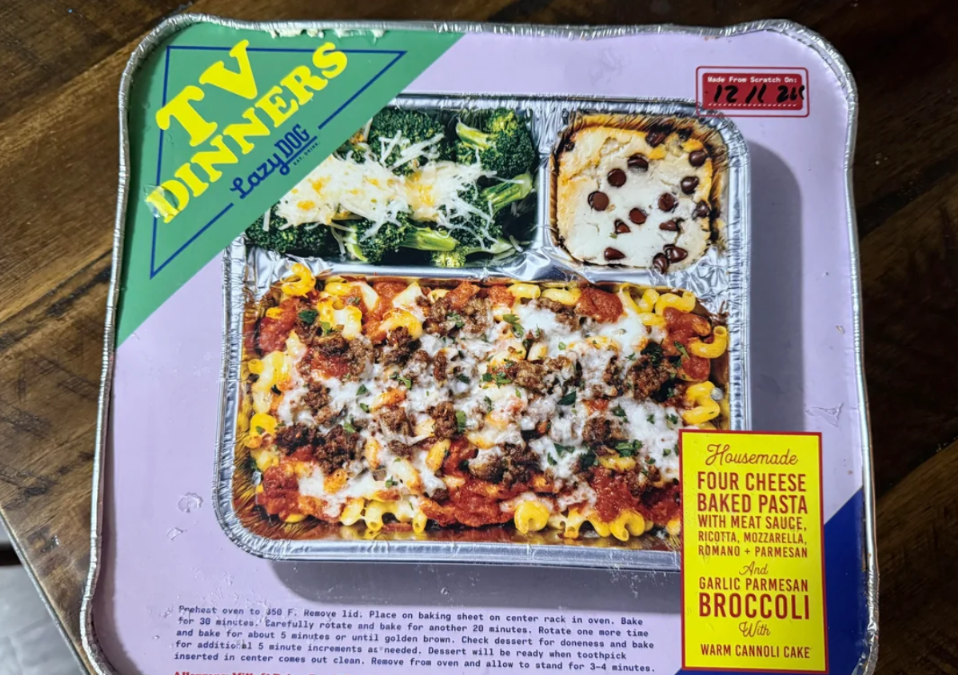 A TV dinner package features a baked pasta with cheese and meat sauce, a side of broccoli with garlic parmesan, and a piece of cannoli cake—reminiscent of nostalgic photos of food items from classic family dinners. The packaging is purple and lists the meal details in a yellow box.