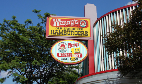 Two Wendy&rsquo;s signs: one reads &ldquo;Wendy&rsquo;s Old Fashioned Hamburgers,&rdquo; and the other says &ldquo;Wendy&rsquo;s 1st Restaurant Est. 1969,&rdquo; with trees and parts of the building visible under a blue sky, hinting at the classic atmosphere where favorite Wendy's menu items first made their debut.
