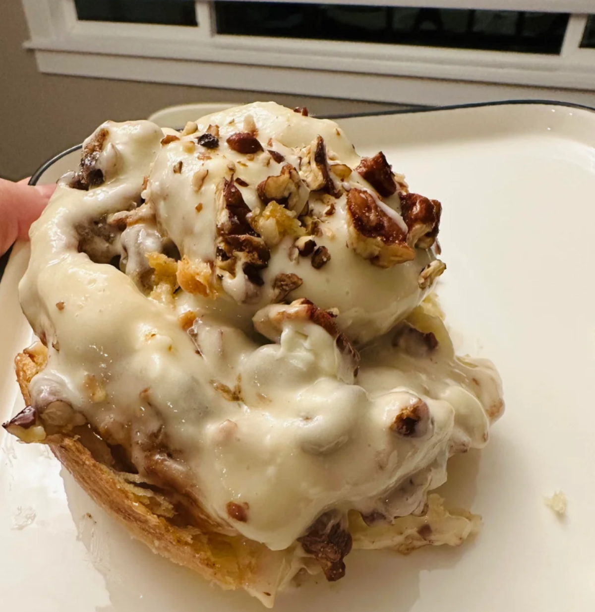 A cinnamon roll topped with thick white icing and chopped pecans, a perfect example of homemade desserts, sits on a white plate in front of a window.