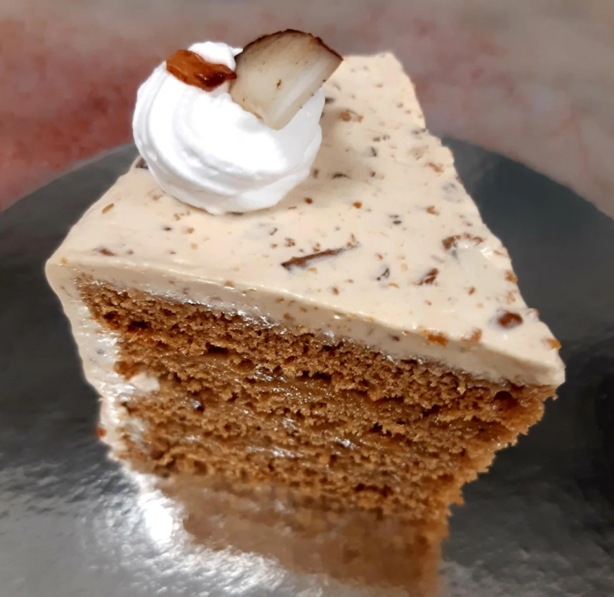 A slice of layered coffee cake with light frosting, chocolate shavings, and a swirl of whipped cream topped with a nut and chocolate piece on a metallic surface—a decadent example of homemade desserts.