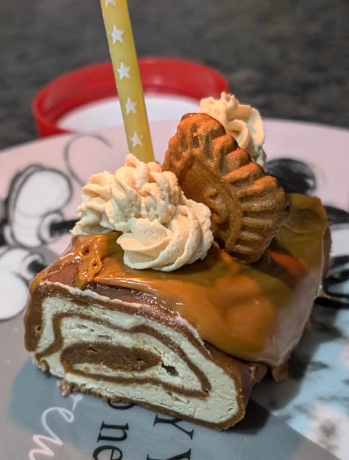 A slice of rolled cake with caramel topping, cream swirls, and two decorative cookies sits on a plate, showcasing the charm of homemade desserts. A yellow straw with white stars is inserted for decoration. The background is slightly blurred.