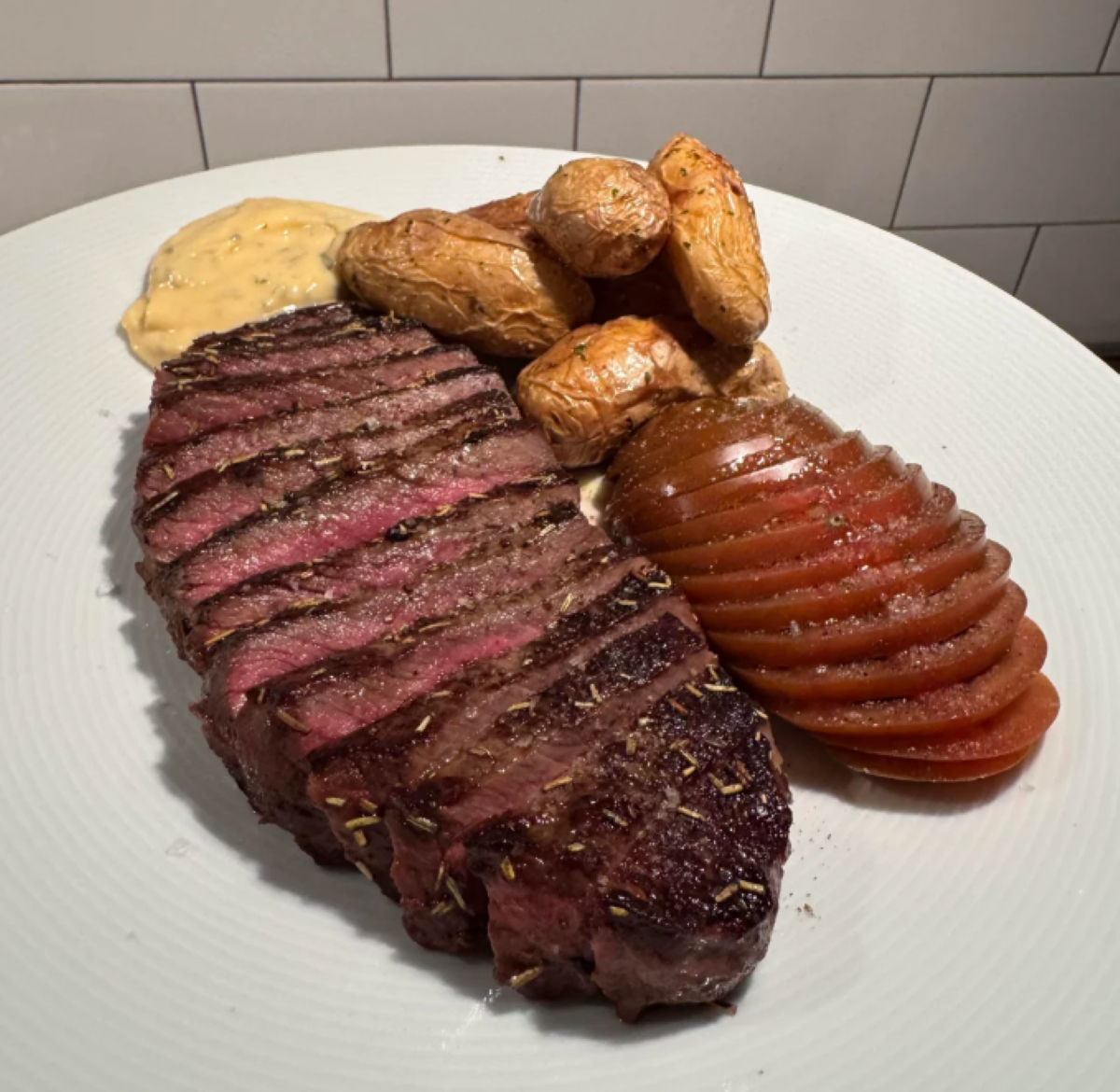A plate with sliced steak, seasoned fingerling potatoes, sliced tomato, and a dollop of creamy sauce, set on a white dish with a tiled background.