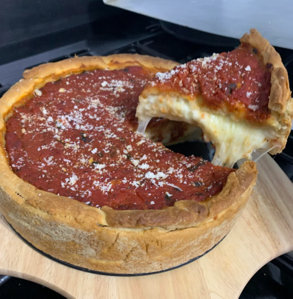 A slice of deep-dish pizza with tomato sauce and melted cheese is being lifted from the whole pizza, which sits on a wooden board.