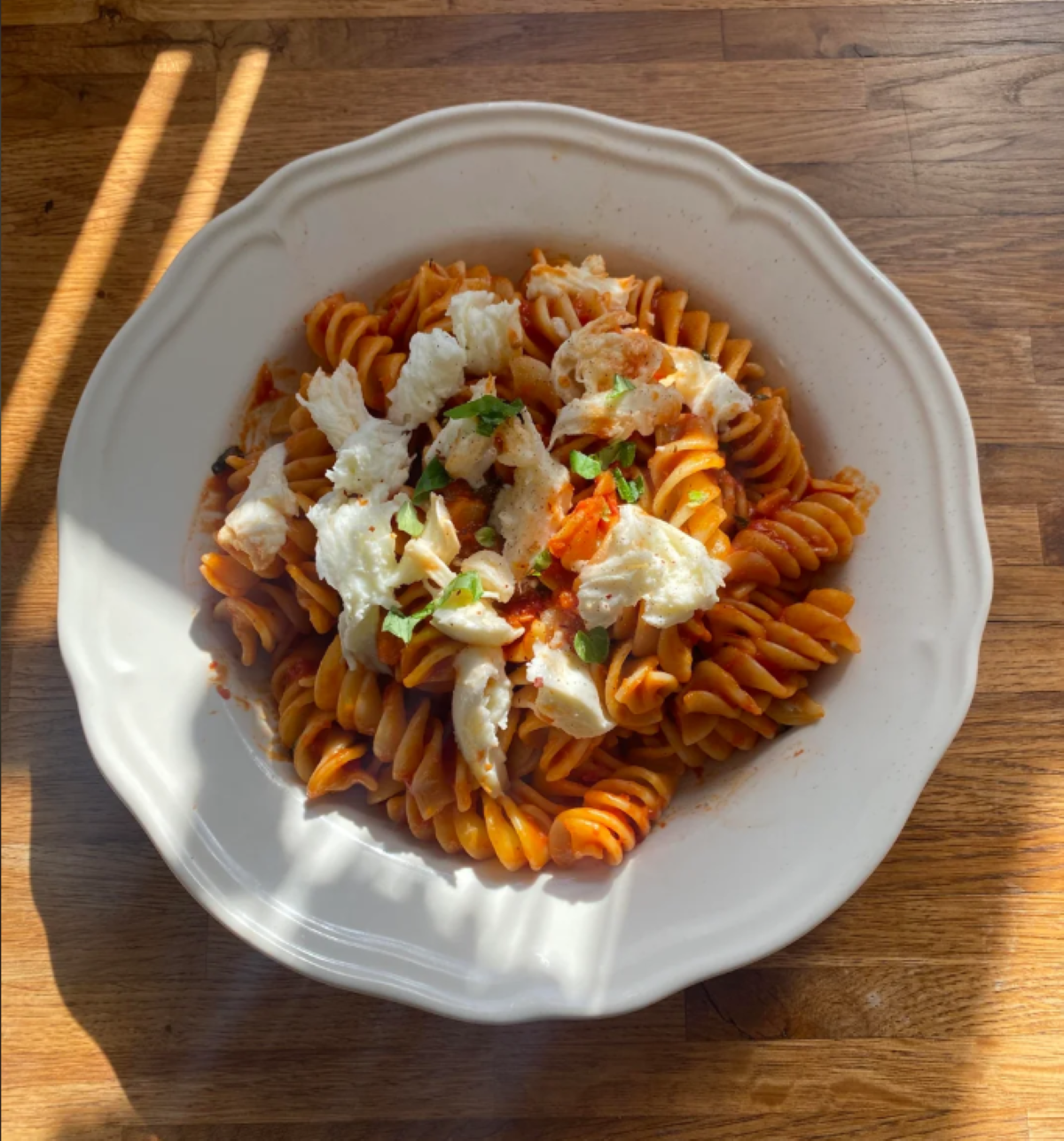A white plate of cooked fusilli pasta with tomato sauce, topped with melted cheese and chopped green herbs, sits on a wooden table in natural sunlight.